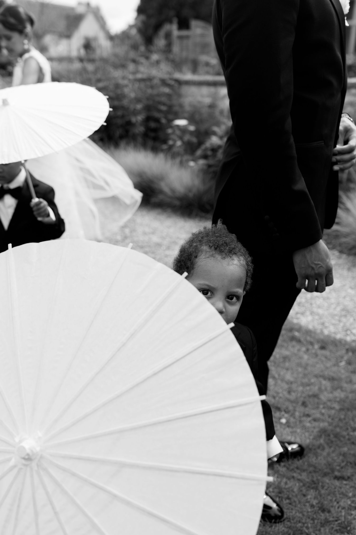 Black and white photo of a young child peeking curiously from behind white parasols at a wedding.