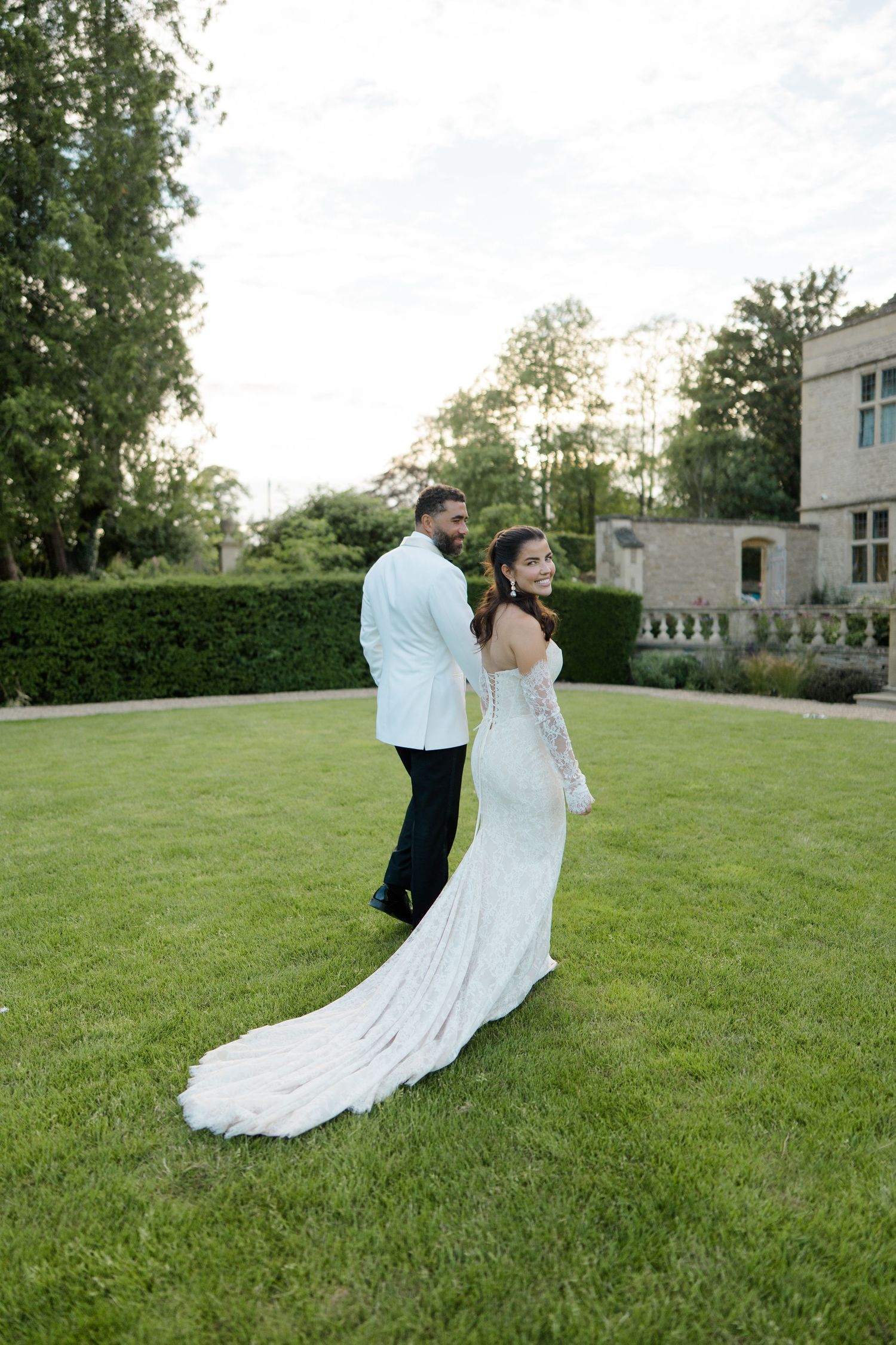 Bride in elegant white gown with long train stands with groom on green lawn before stone manor.