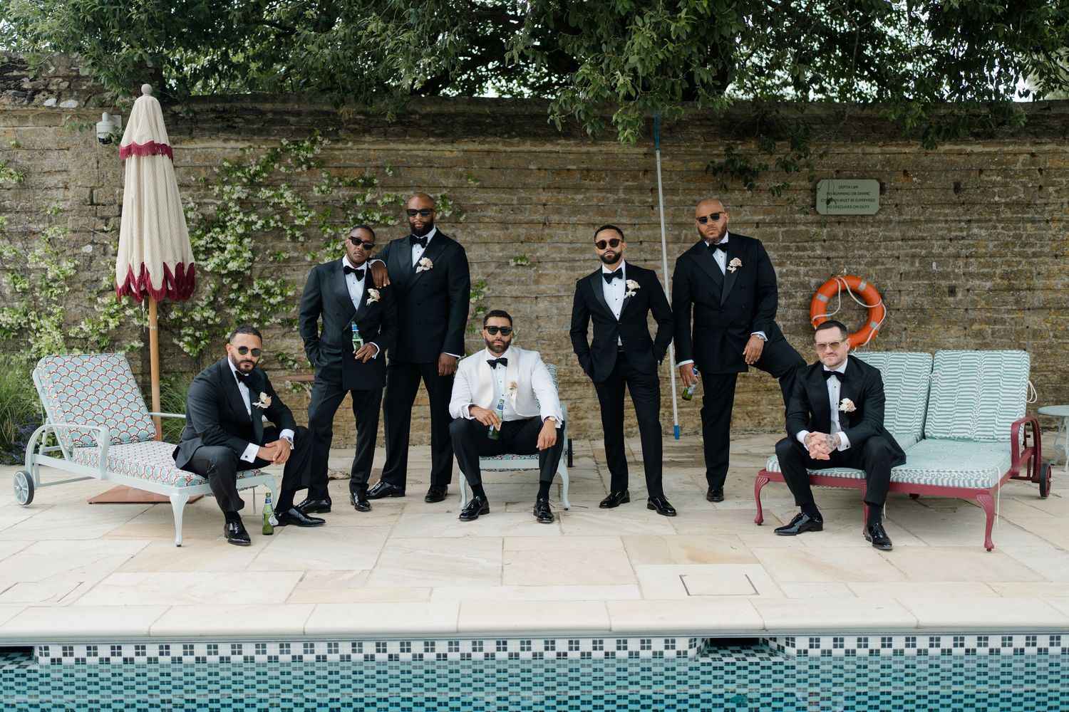 Groomsmen in black tuxedos pose poolside at elegant wedding venue on sunny day.