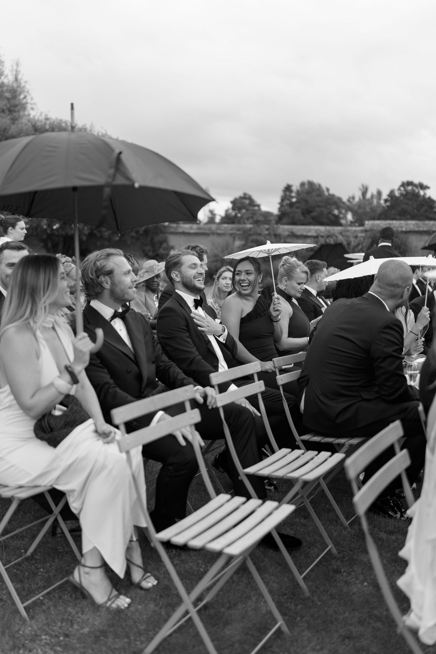 Wedding guests laughing and sheltering under umbrellas during an outdoor ceremony on a cloudy day.