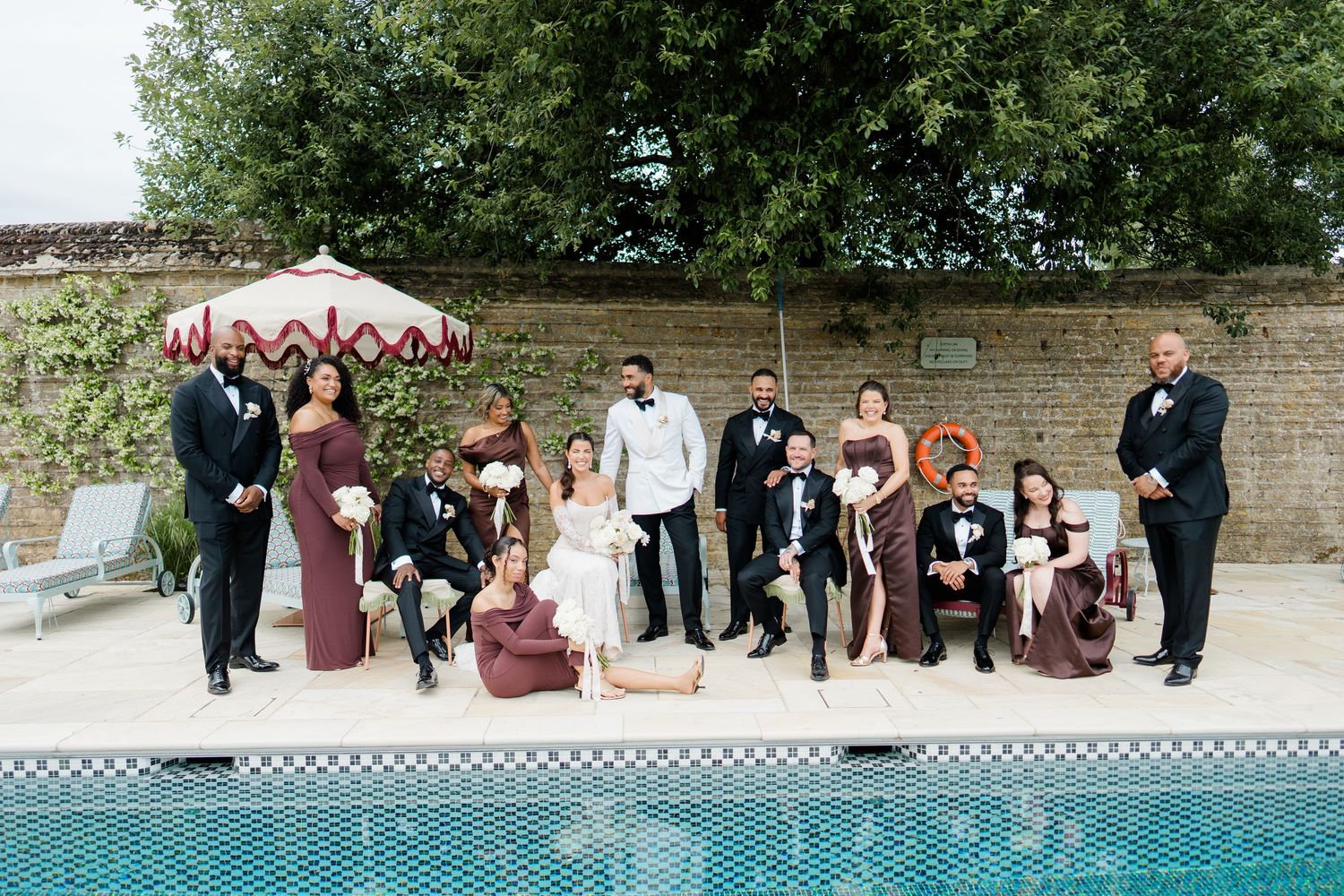 Wedding party poses poolside in burgundy and black formal wear at elegant outdoor venue.