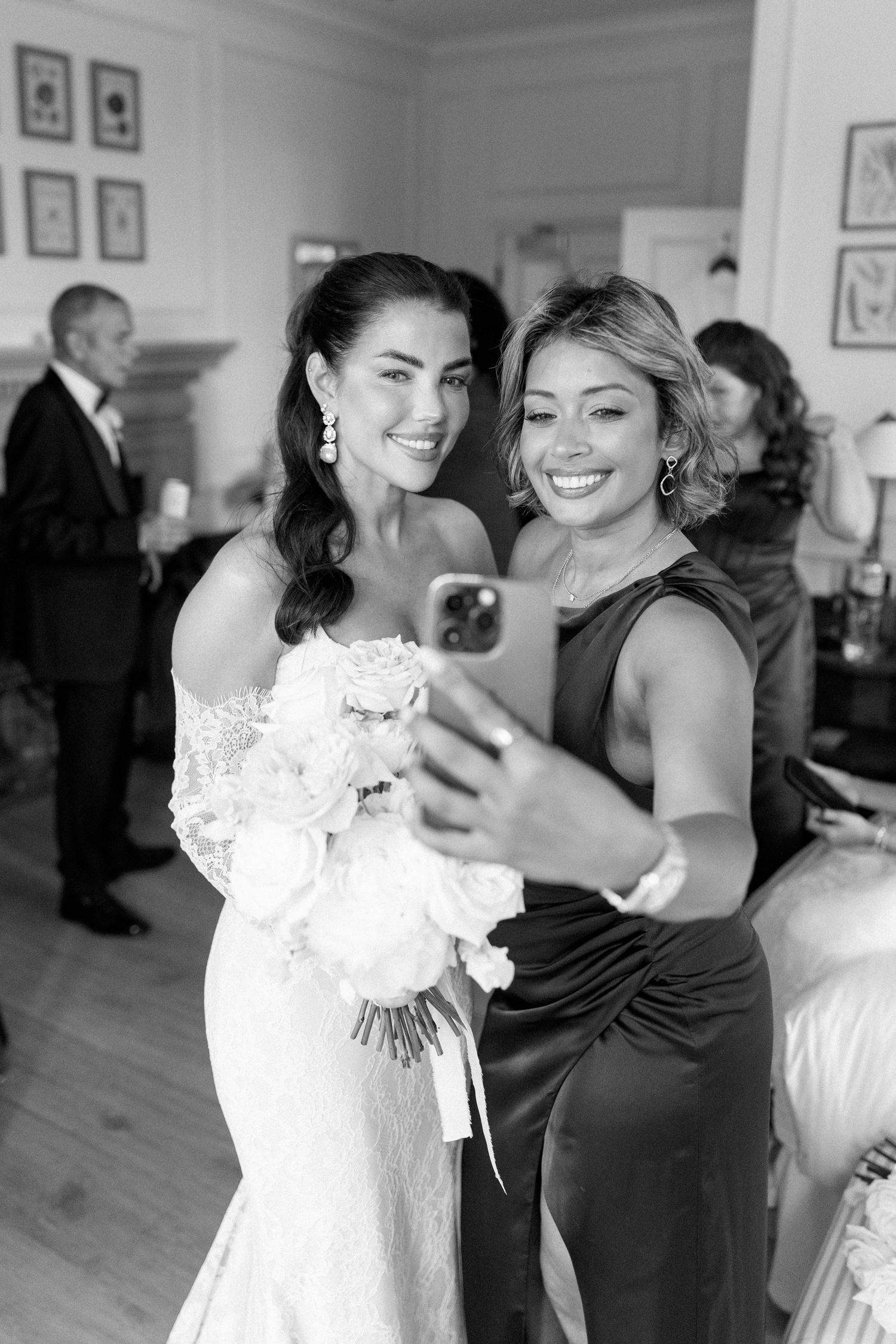 Bride in white gown holding bouquet taking selfie with bridesmaid in black dress, black and white.