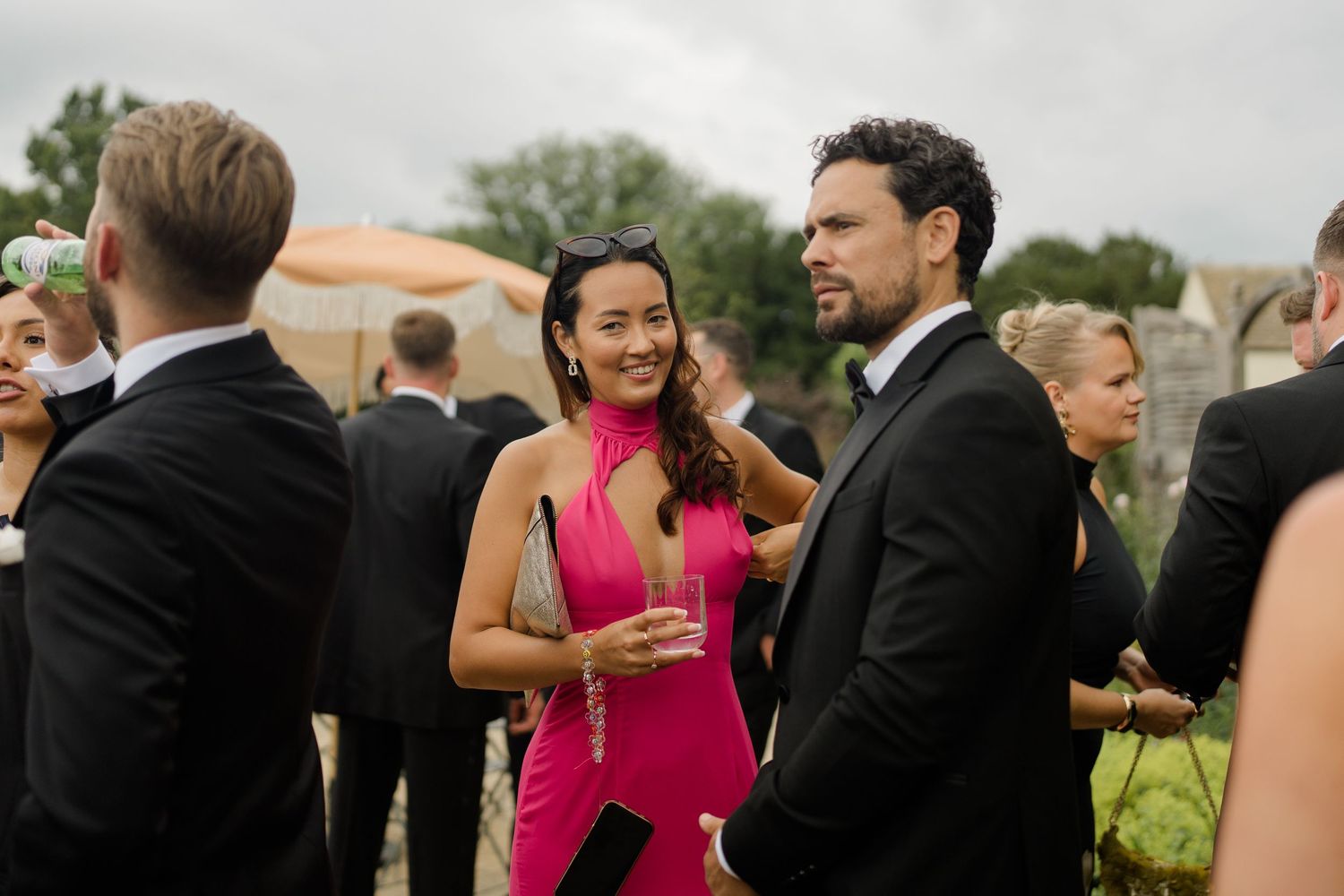 Wedding guest in pink halter dress laughing with man in black suit at outdoor reception.