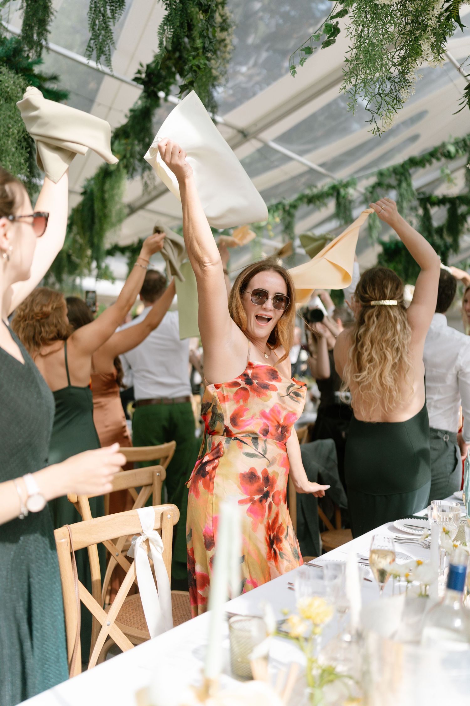 Wedding reception guests celebrating with raised napkins under a greenhouse-style venue with hanging greenery.