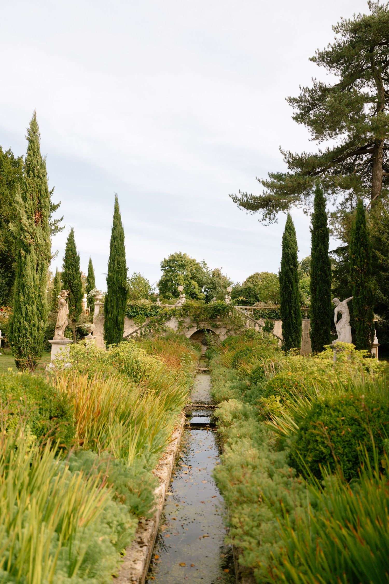 Formal garden path lined with tall cypress trees and ornamental grasses leading to water feature.