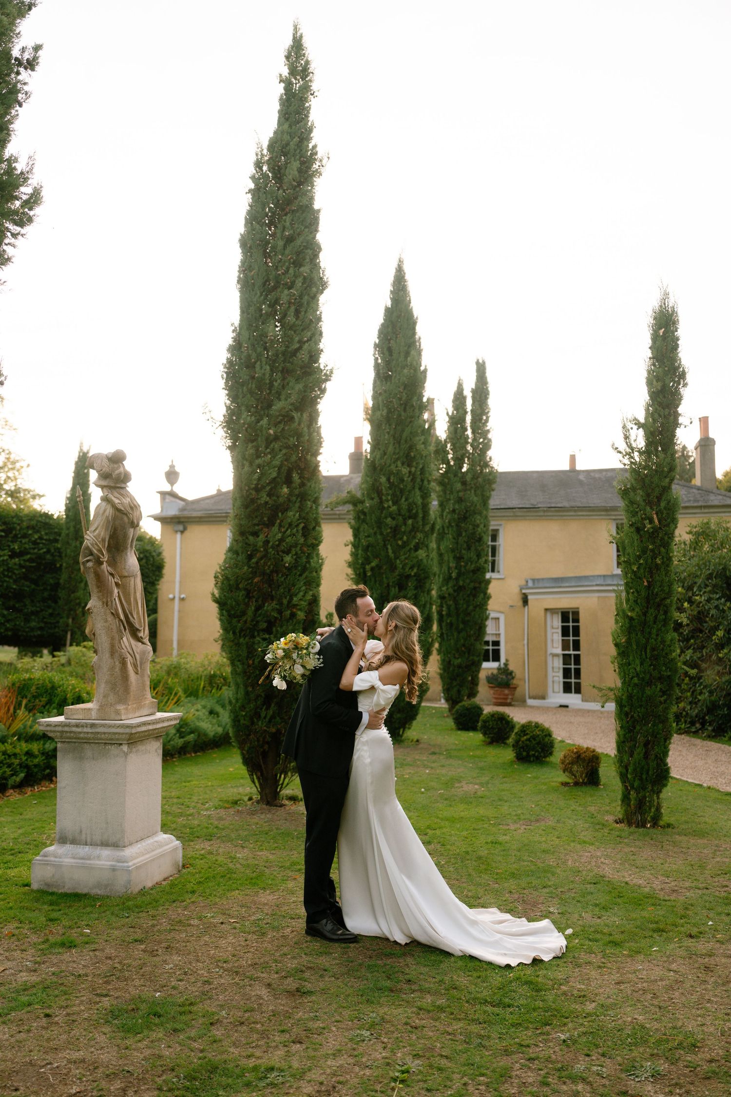 A wedding couple embraces in an elegant garden with classical statues and cypress trees.