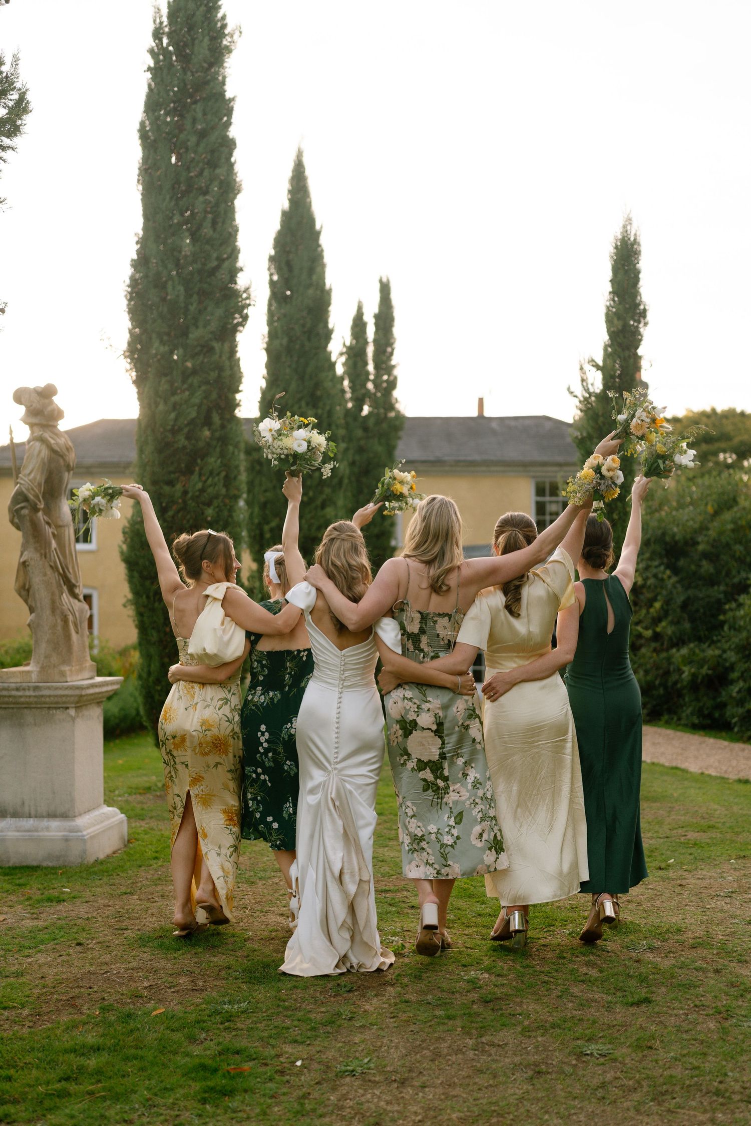 Wedding party members holding hands and celebrating with raised arms on garden grounds.