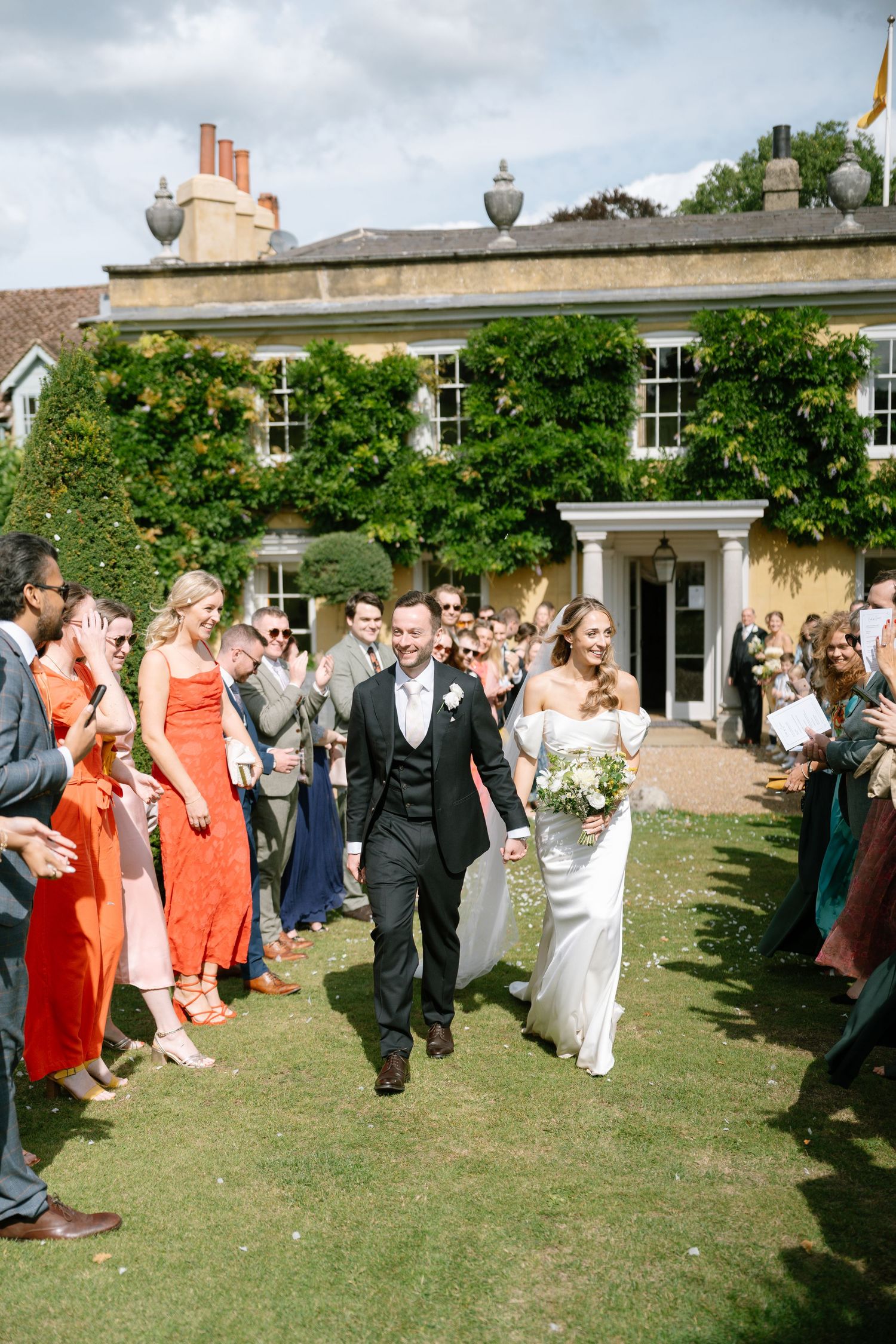 Wedding party walks through a corridor of guests on a grassy lawn outside a historic building.
