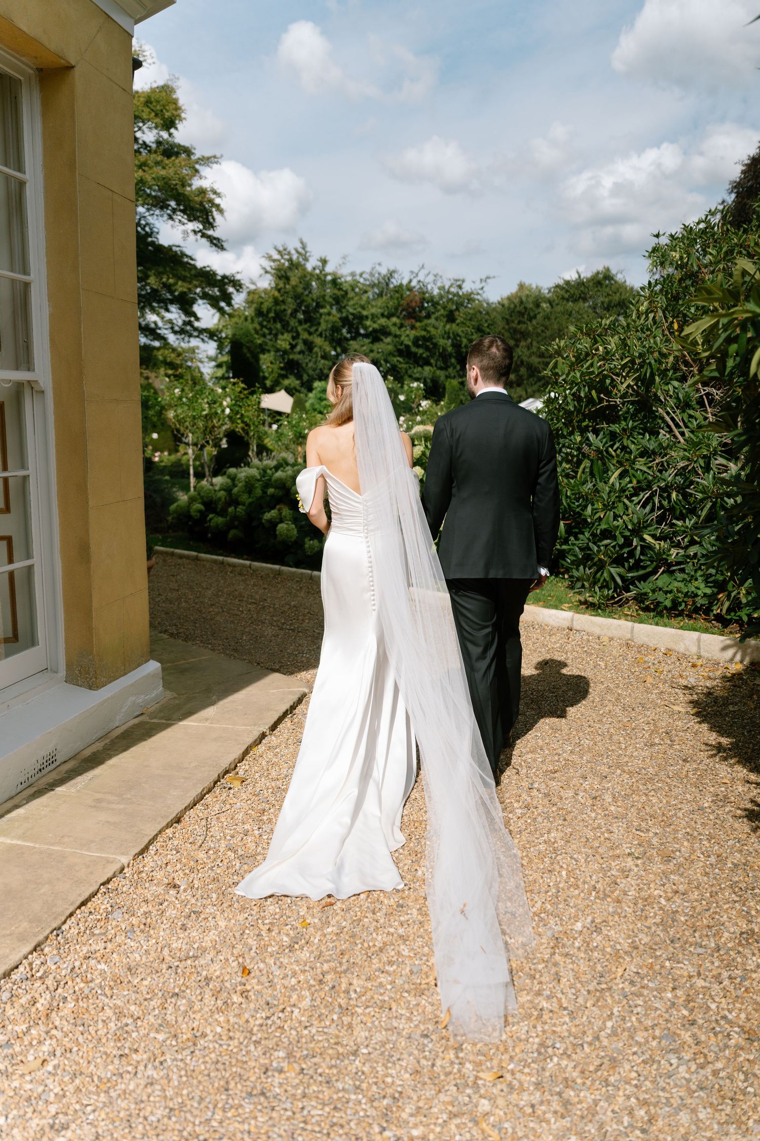 Wedding couple walks along gravel path beside yellow building with flowing veil and formal attire.