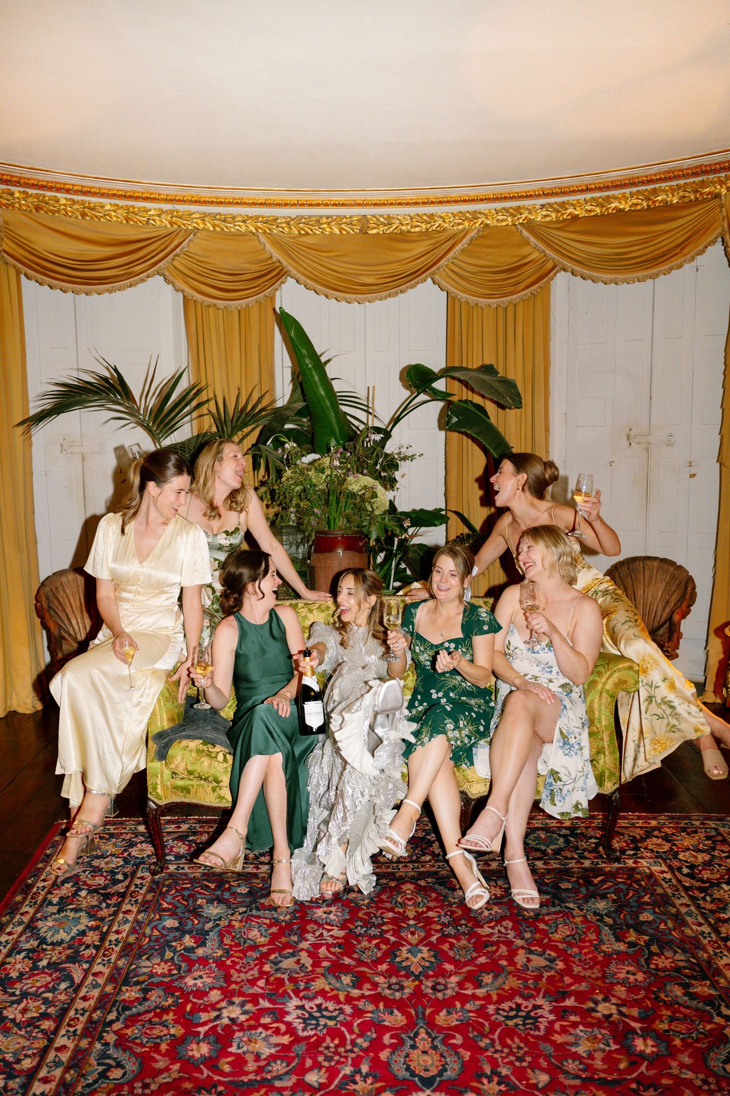 Wedding party relaxing on plush furniture atop a red patterned carpet in an ornate room.