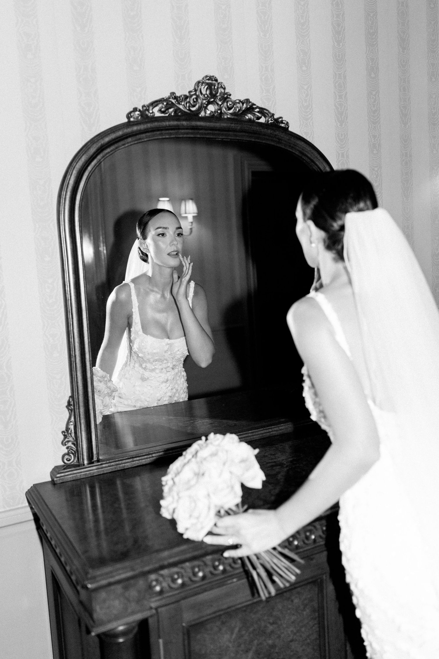 Bride in white lace dress looks at reflection in antique mirror during wedding preparations.