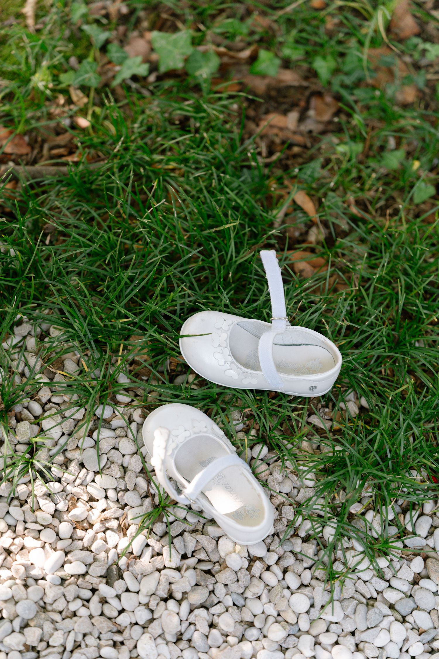 White baby shoes arranged on decorative white pebbles in grass.