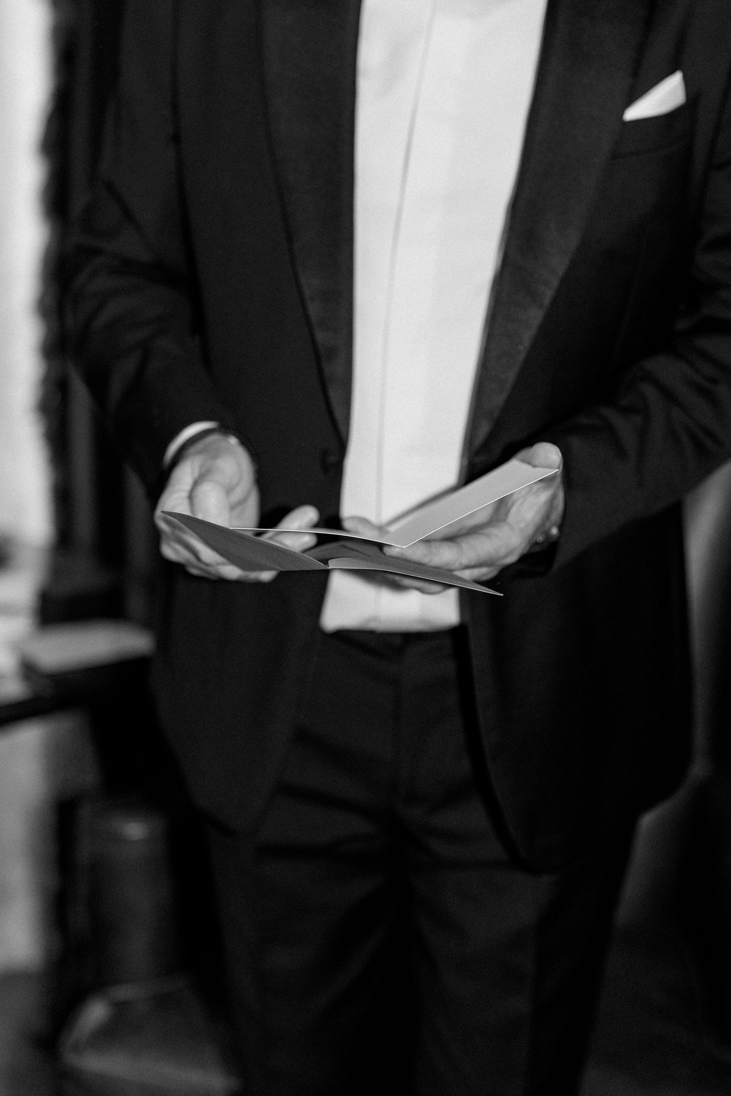 Close-up of hands holding papers while wearing a formal black tuxedo and white shirt.