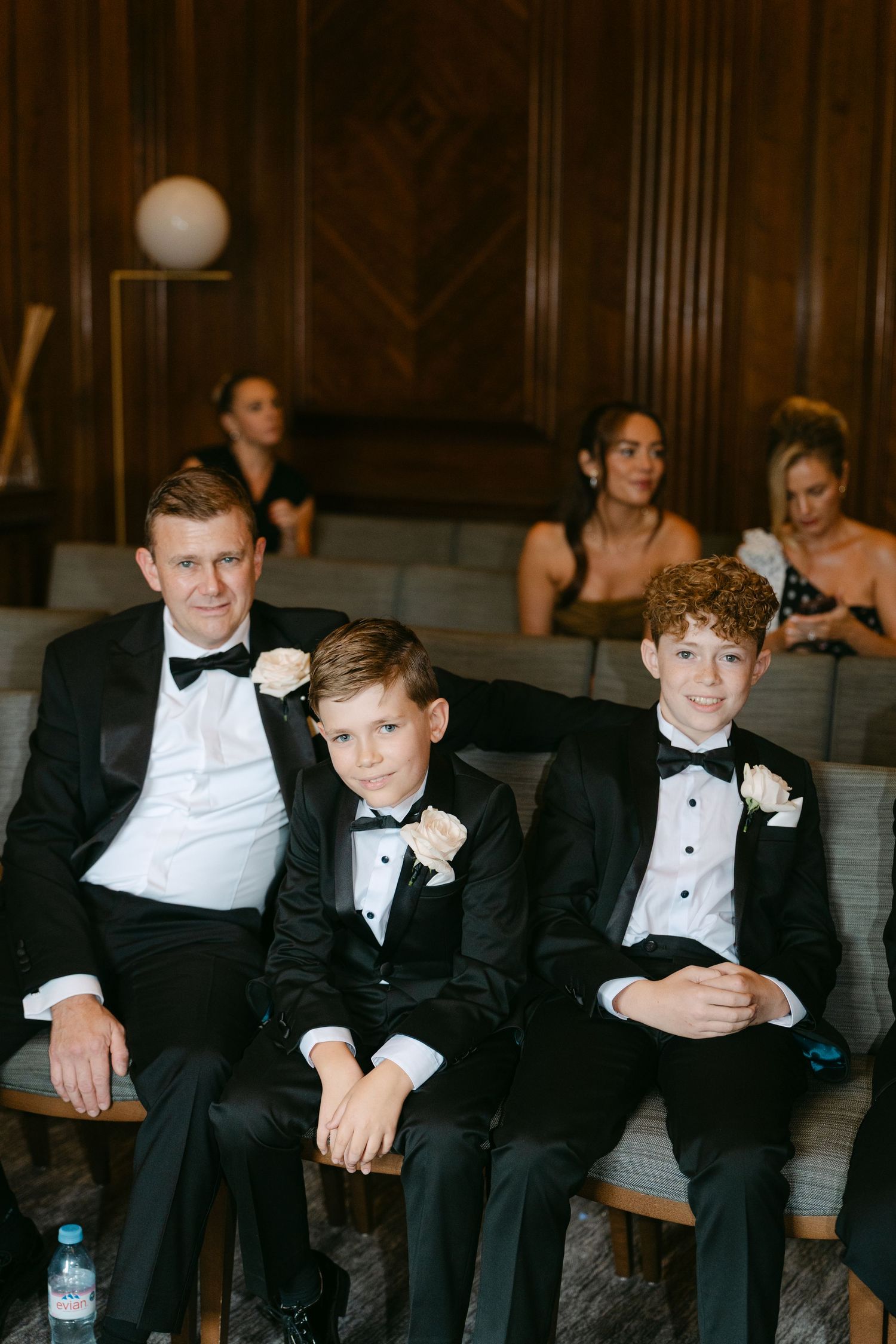 Formal wedding party members in tuxedos seated on wooden benches.