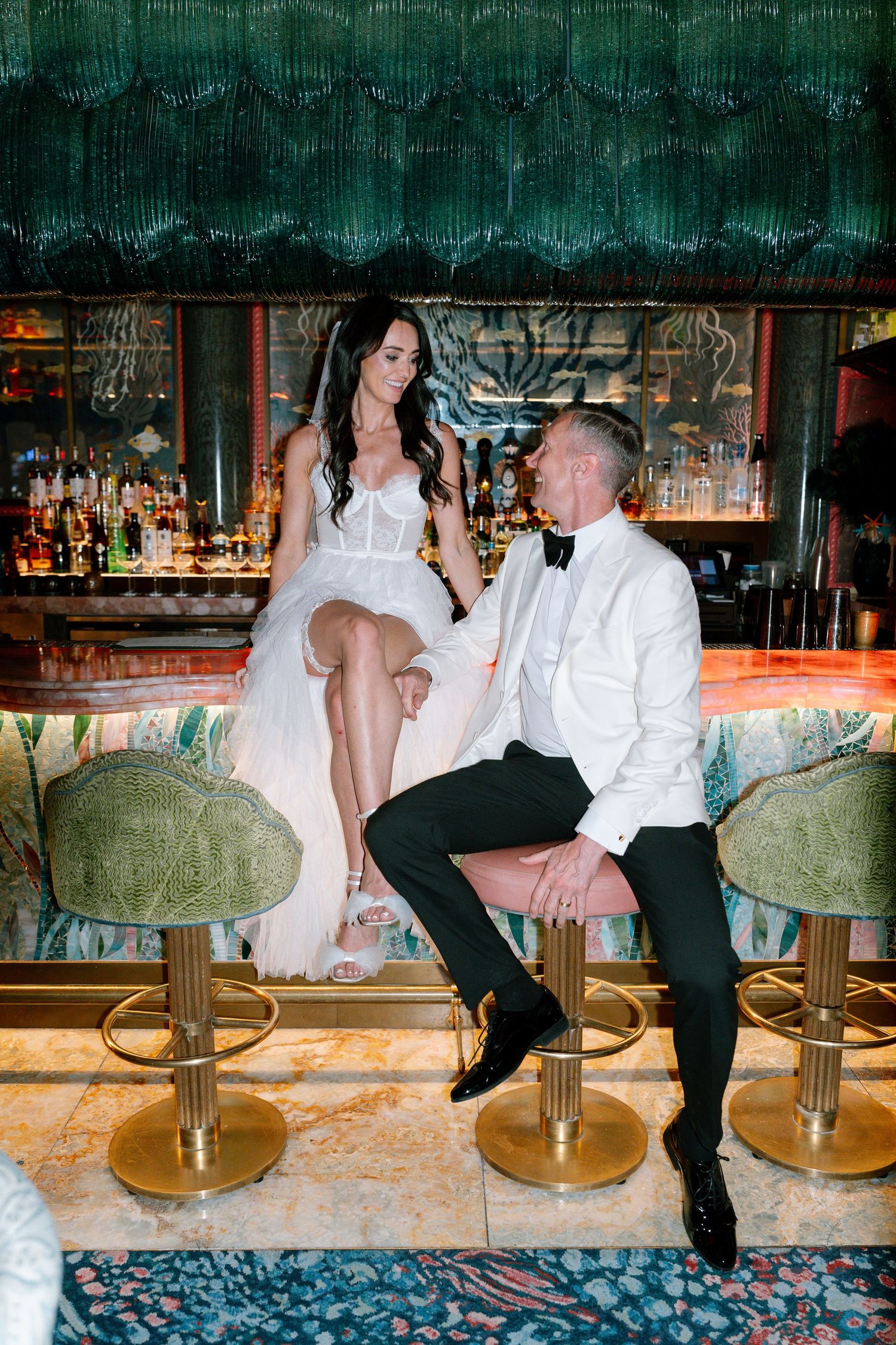 A couple in formal attire sits playfully at a bar counter with retro gold bar stools.