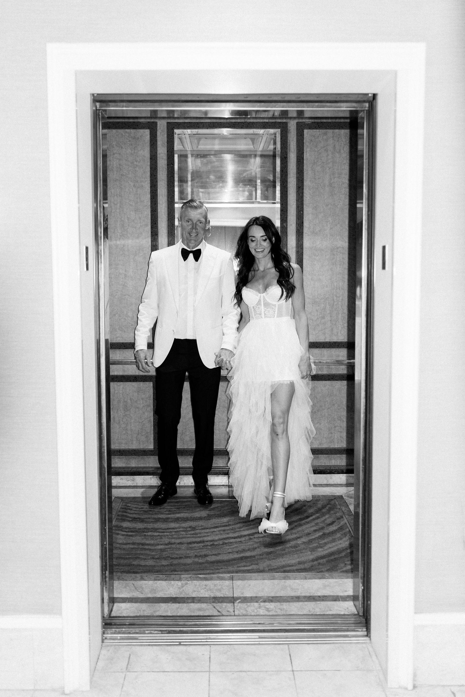A couple in wedding attire stands in an elevator, with the bride wearing a flowing white dress and groom in white tuxedo.