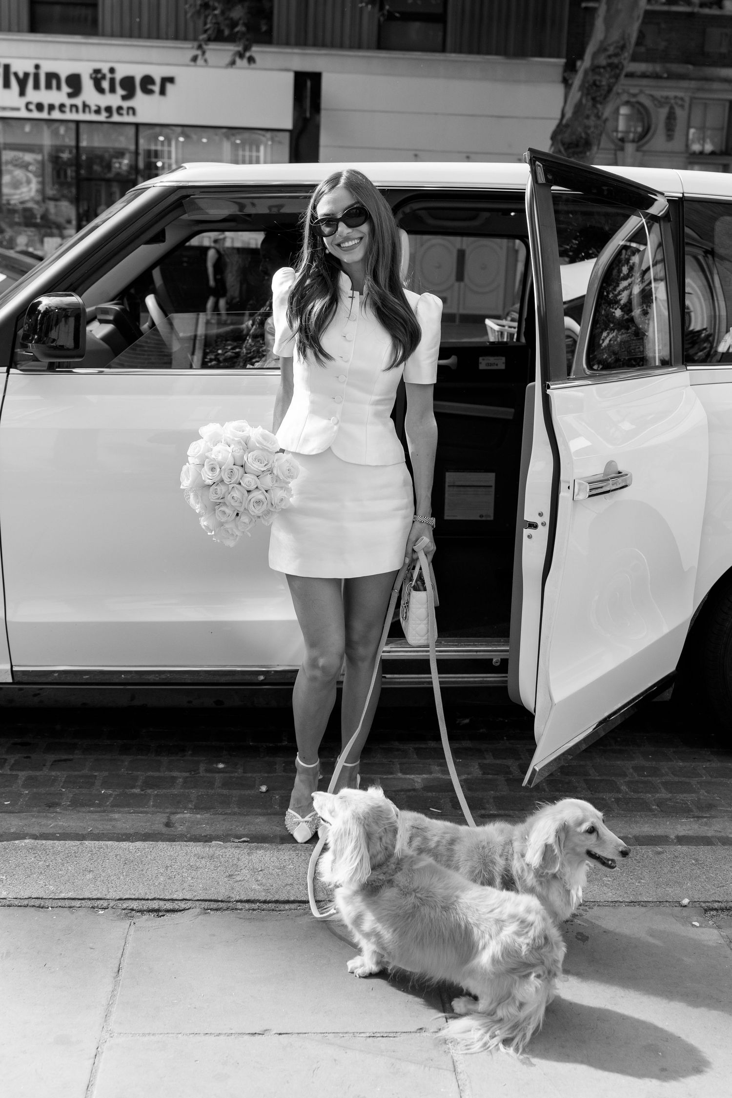 Bride in white mini dress holds rose bouquet with two dogs on leash beside a white taxi cab.