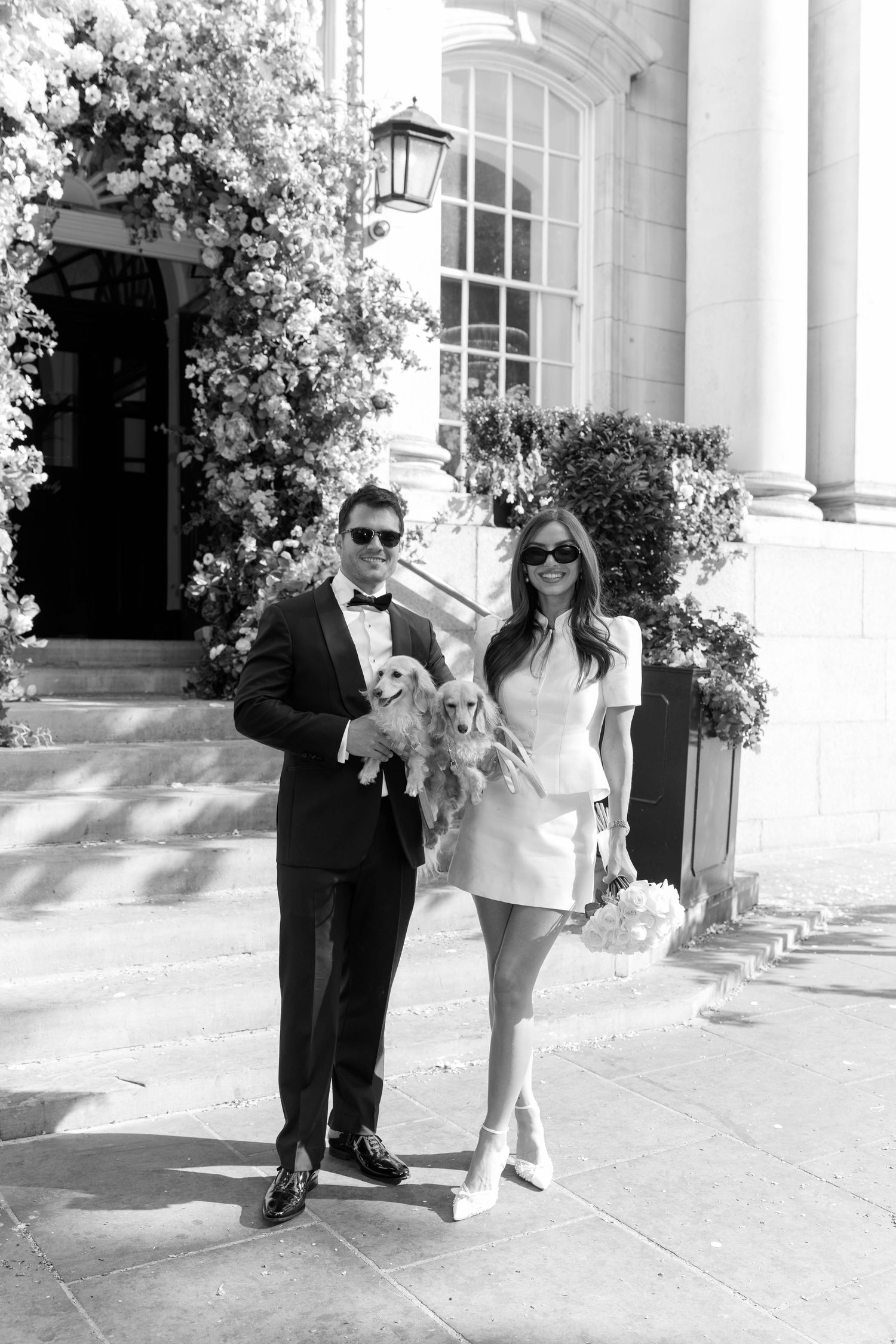 Black and white photo of bride and groom in tuxedo holding a Cocker Spaniel outside elegant venue entrance.