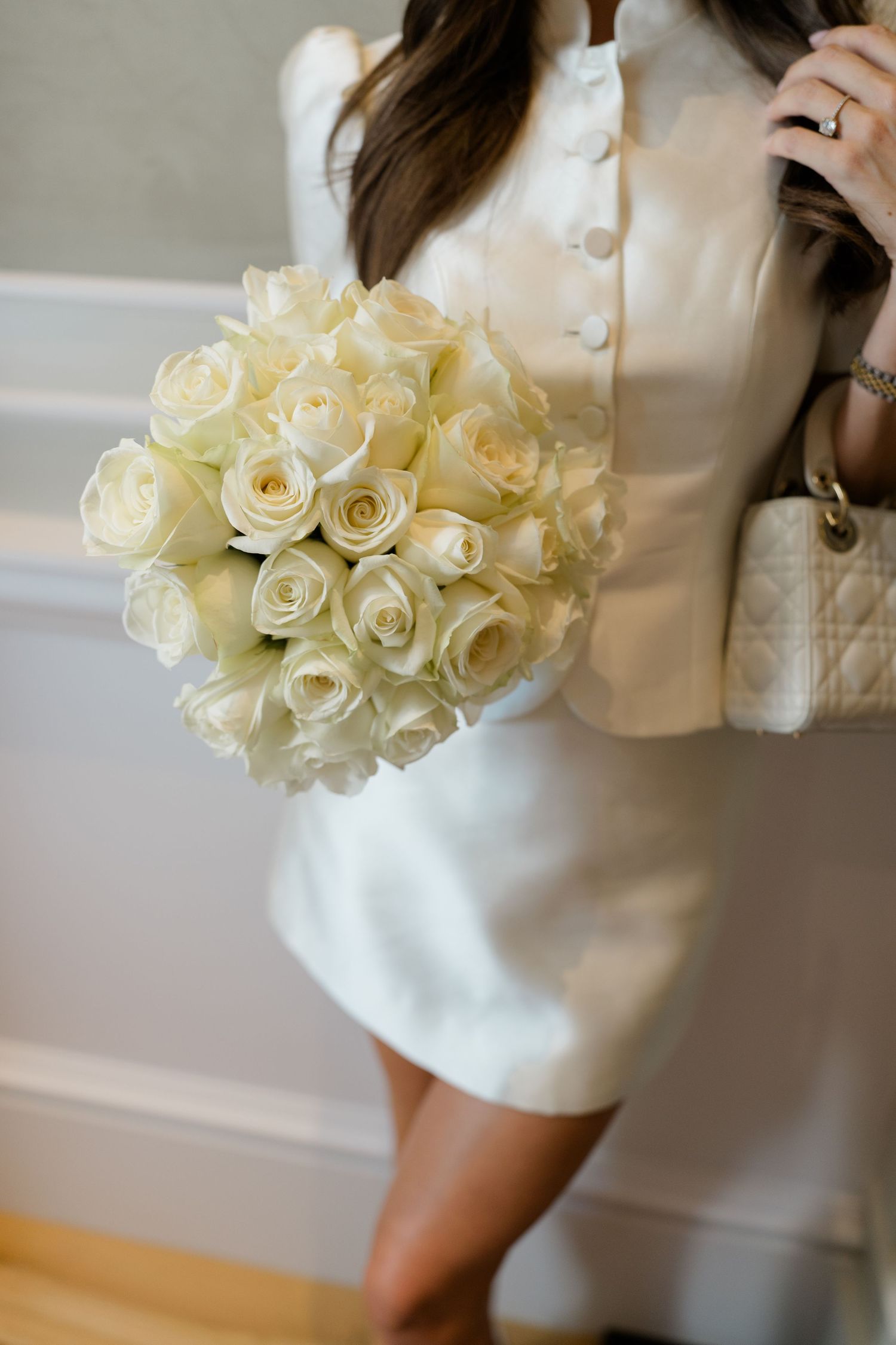 Bride in white mini dress and jacket holds a round bouquet of white roses with a Lady Dior bag.