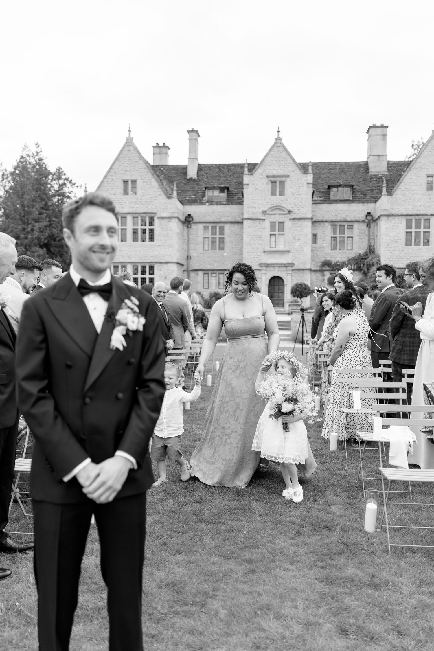 Groom in black tuxedo smiles as bridesmaid walks flower girl down outdoor aisle at elegant manor wedding.