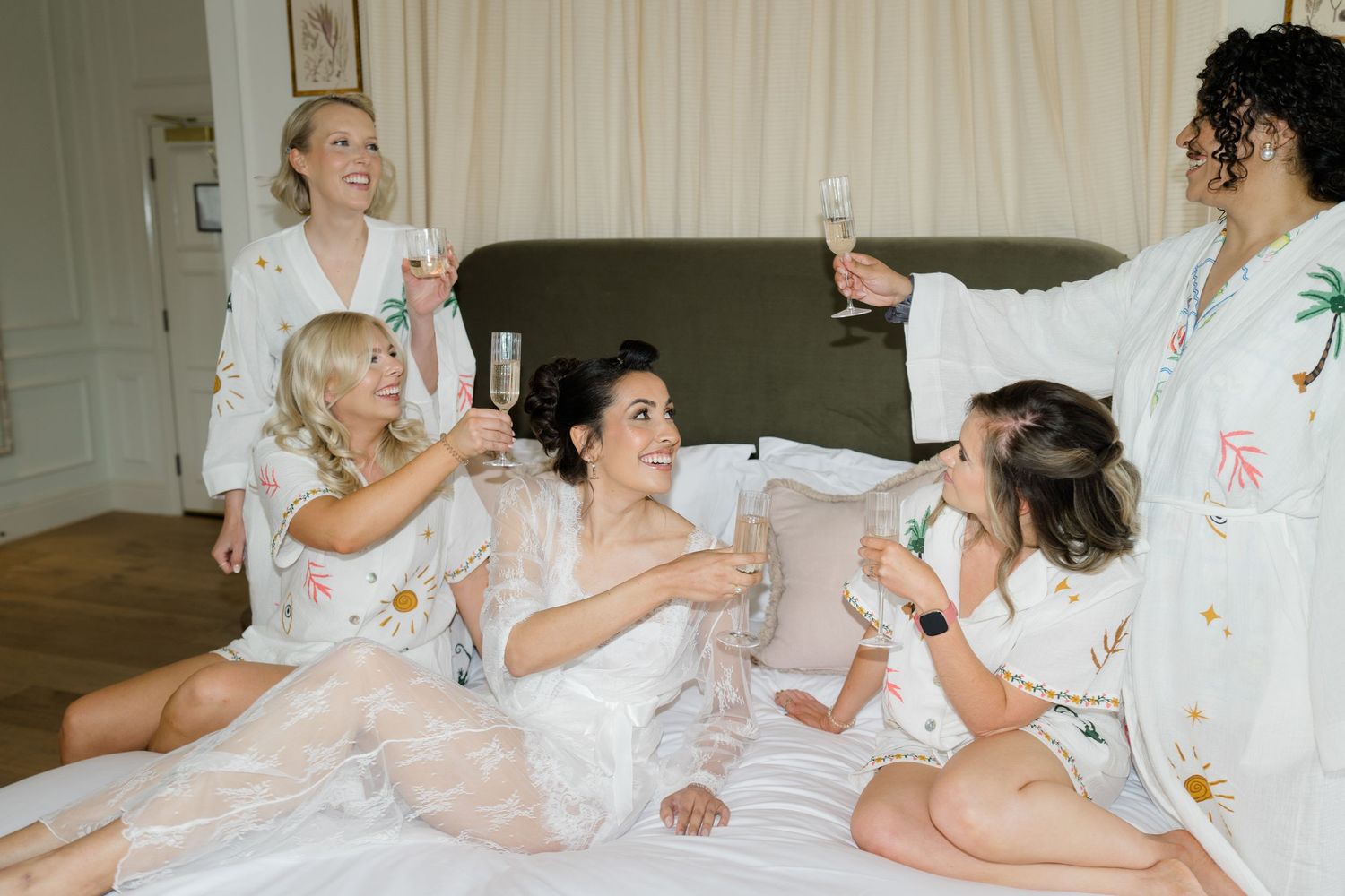 Bridesmaids in floral robes toasting champagne with bride on a bed during wedding morning preparations.