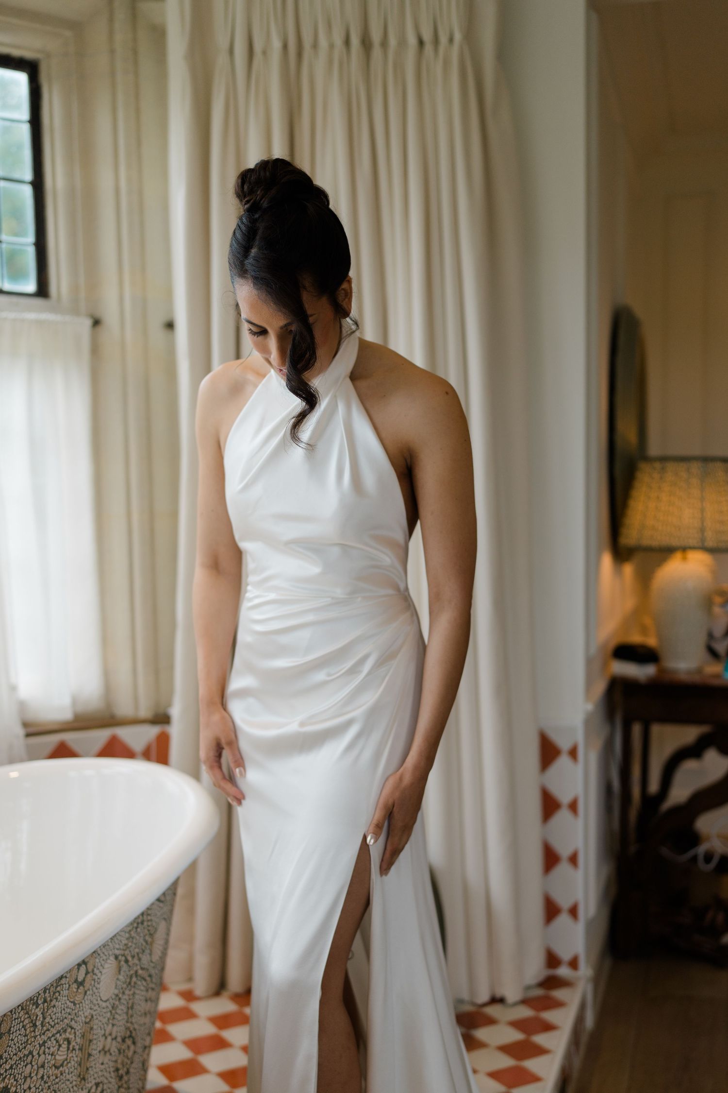 Bride in elegant white halter neck wedding gown standing beside a clawfoot tub on a checkered tile floor.
