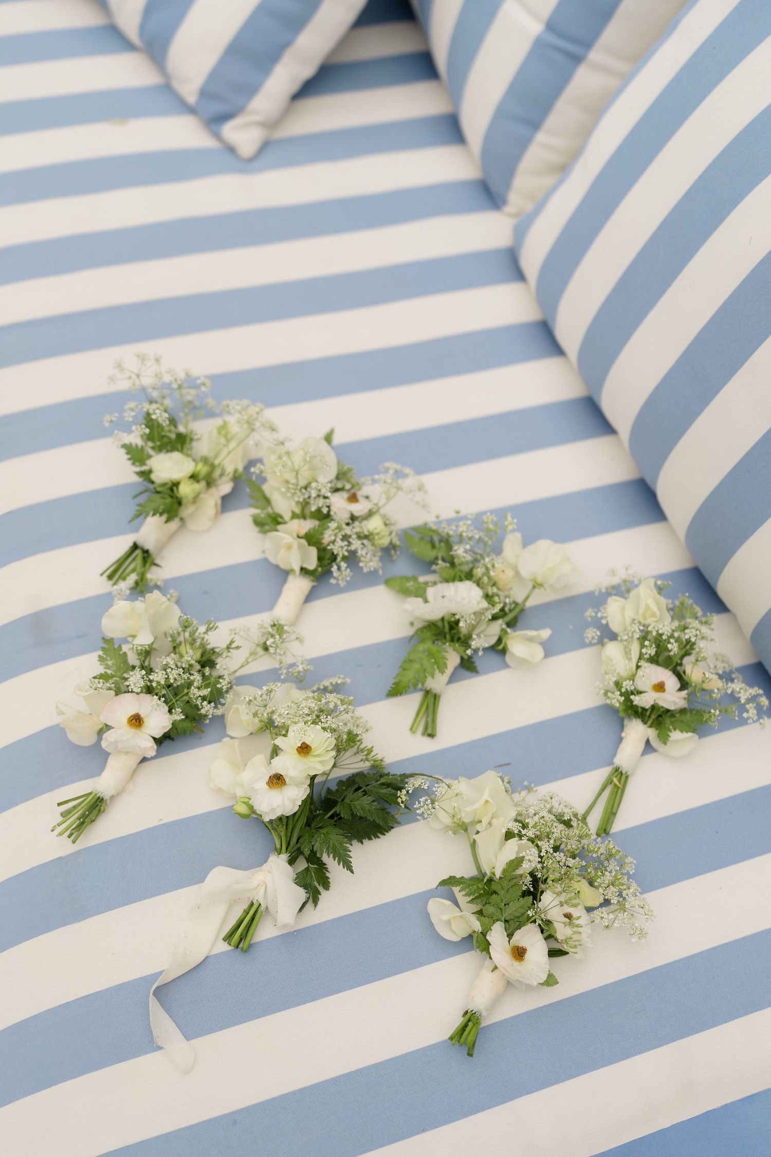 Six small white wedding boutonnieres with baby's breath arranged on blue and white striped fabric.