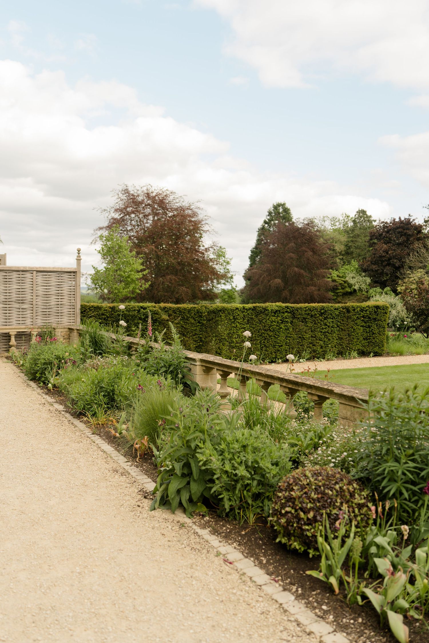 A formal English garden path with herbaceous borders, neatly trimmed hedges, and mature trees beyond.