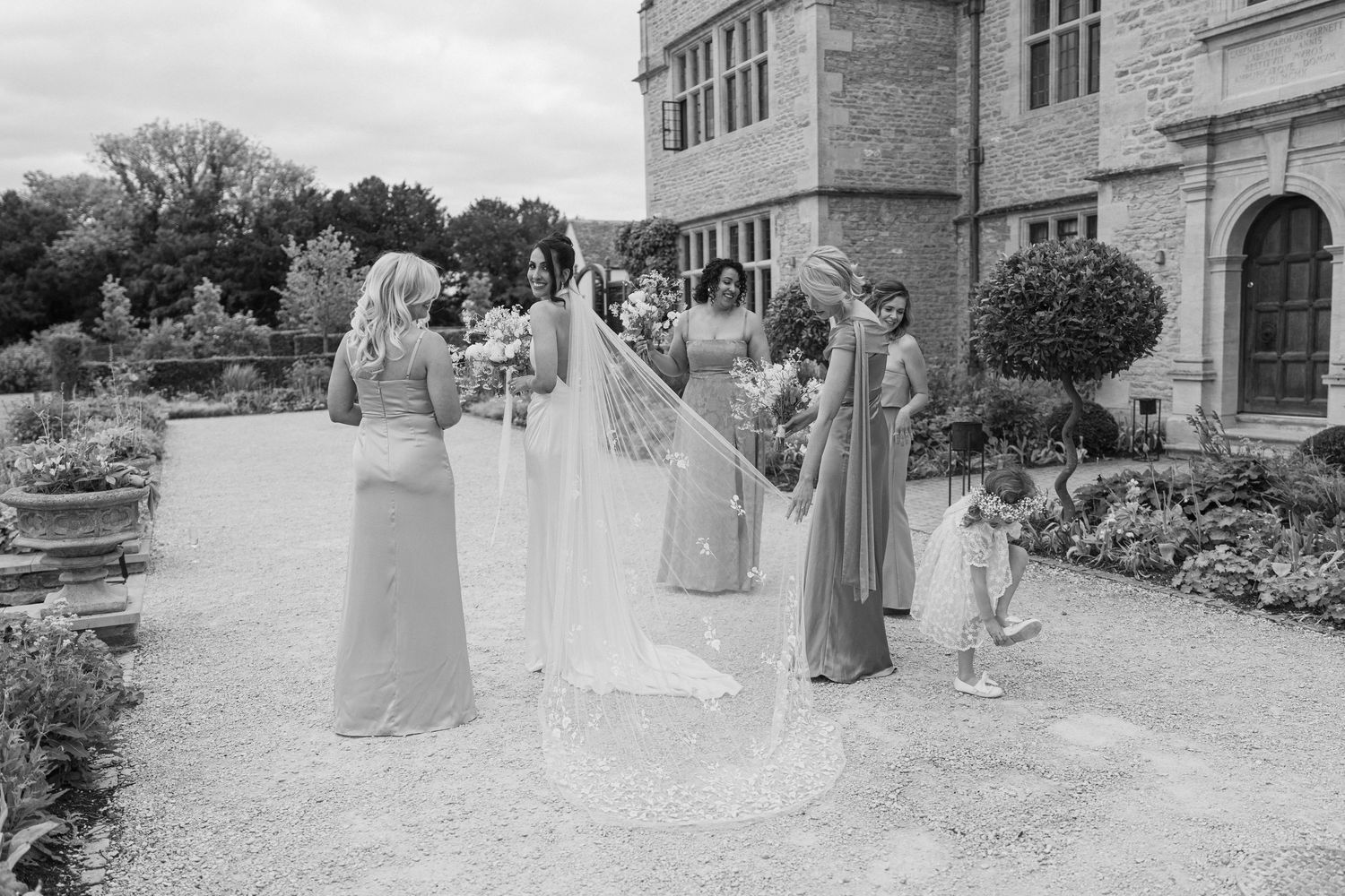 Black and white photo of bridesmaids arranging bride's veil outside historic stone manor wedding venue.