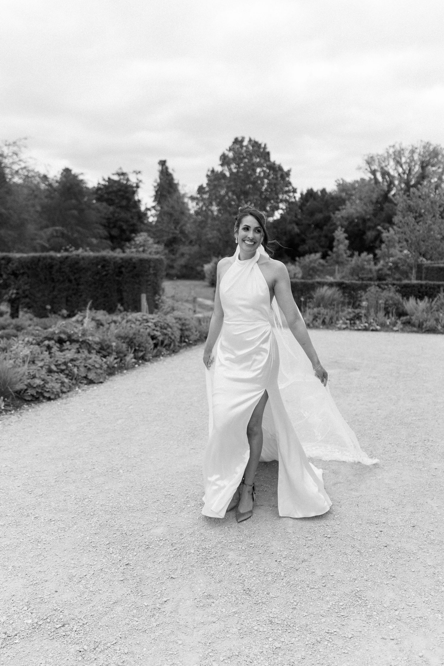 Bride in elegant halter neck white gown with thigh split walks through formal garden in black and white.