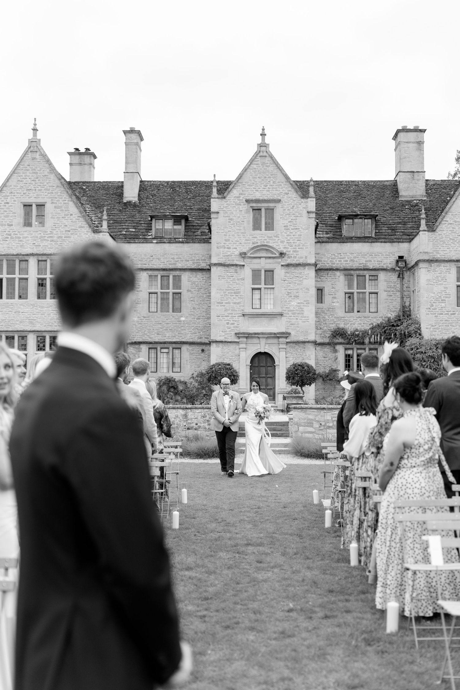 Black and white outdoor wedding ceremony with bride walking down aisle toward groom at historic manor house.