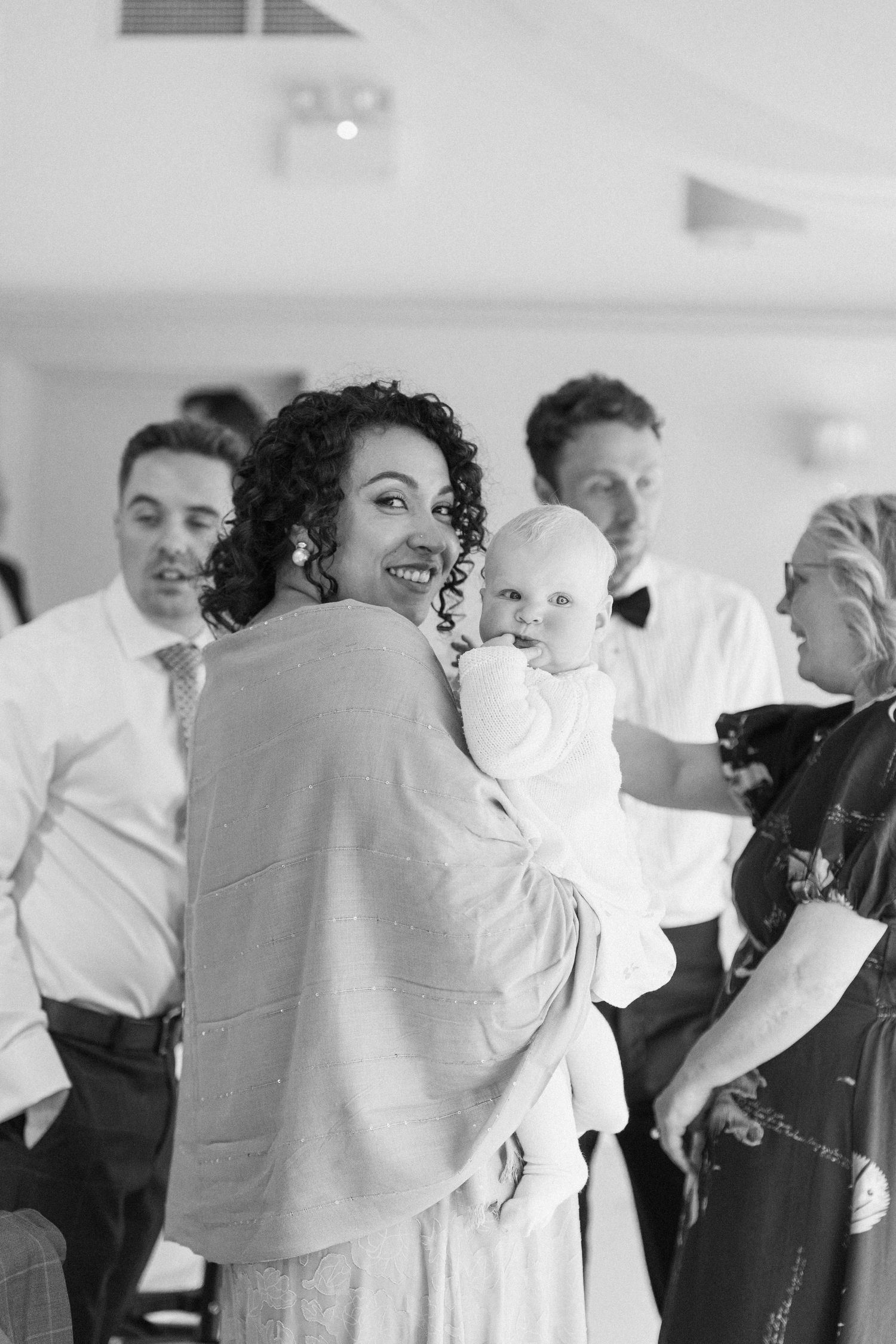 Black and white photo of smiling woman holding baby at a wedding reception surrounded by guests.