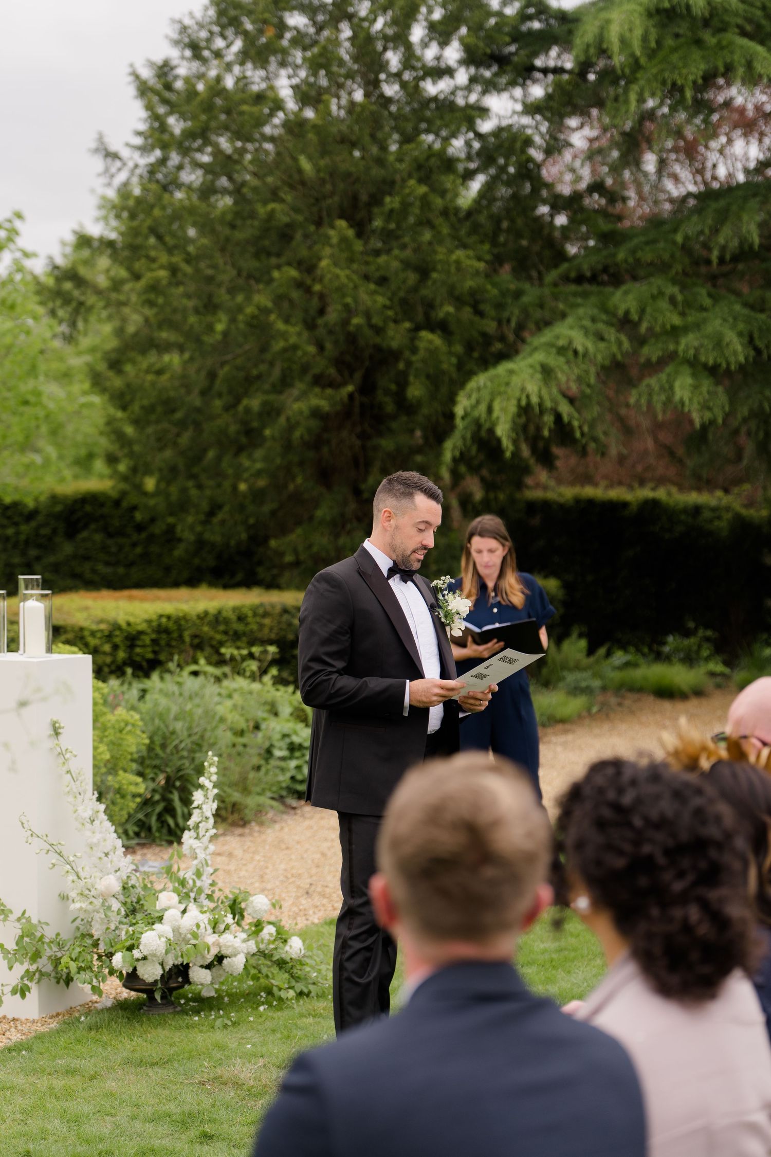 Officiant reads from book during elegant outdoor garden wedding ceremony with guests seated on lawn.