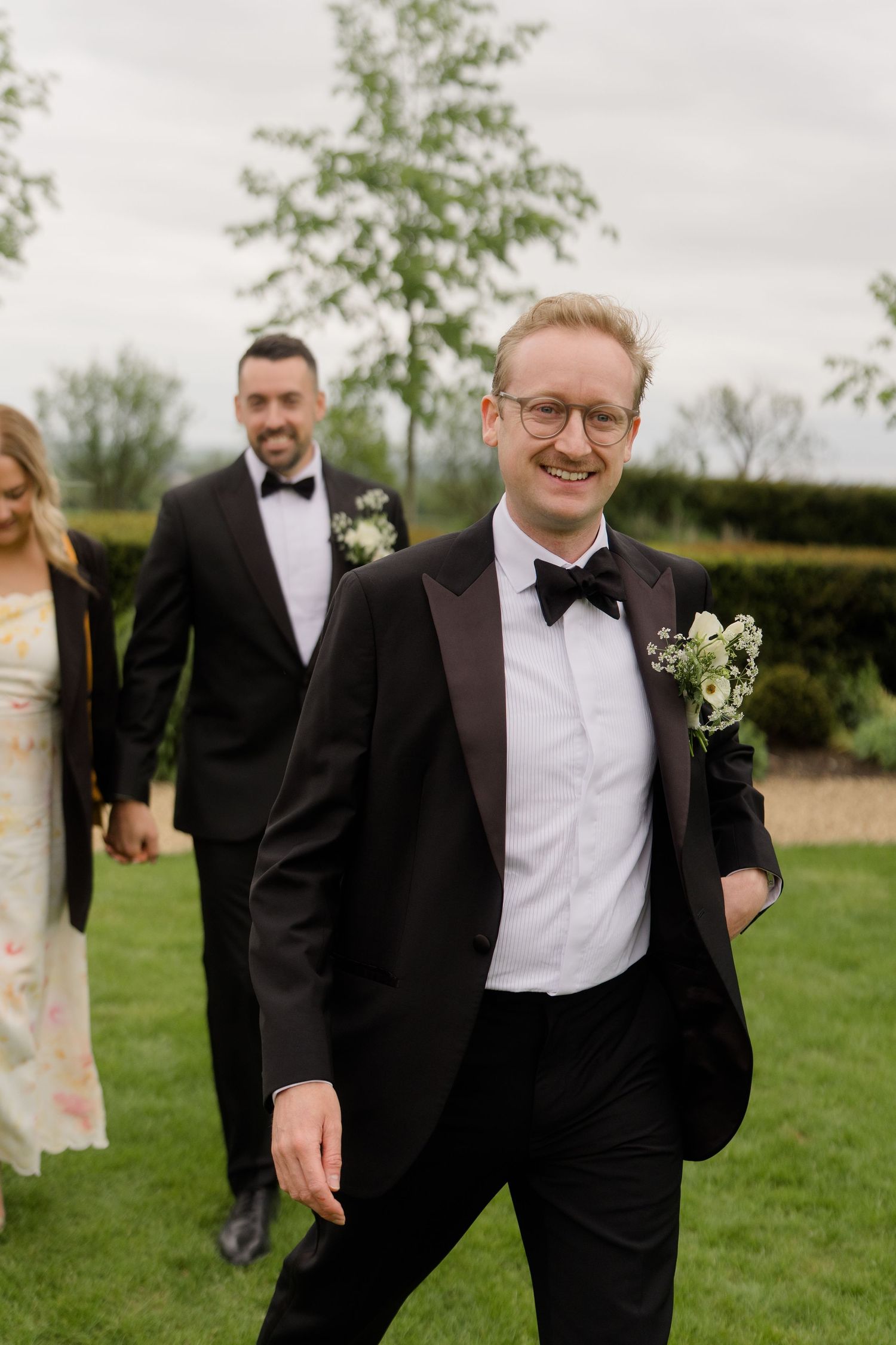 Smiling groom in black tuxedo with boutonniere walks outdoors at wedding venue with groomsmen behind him.