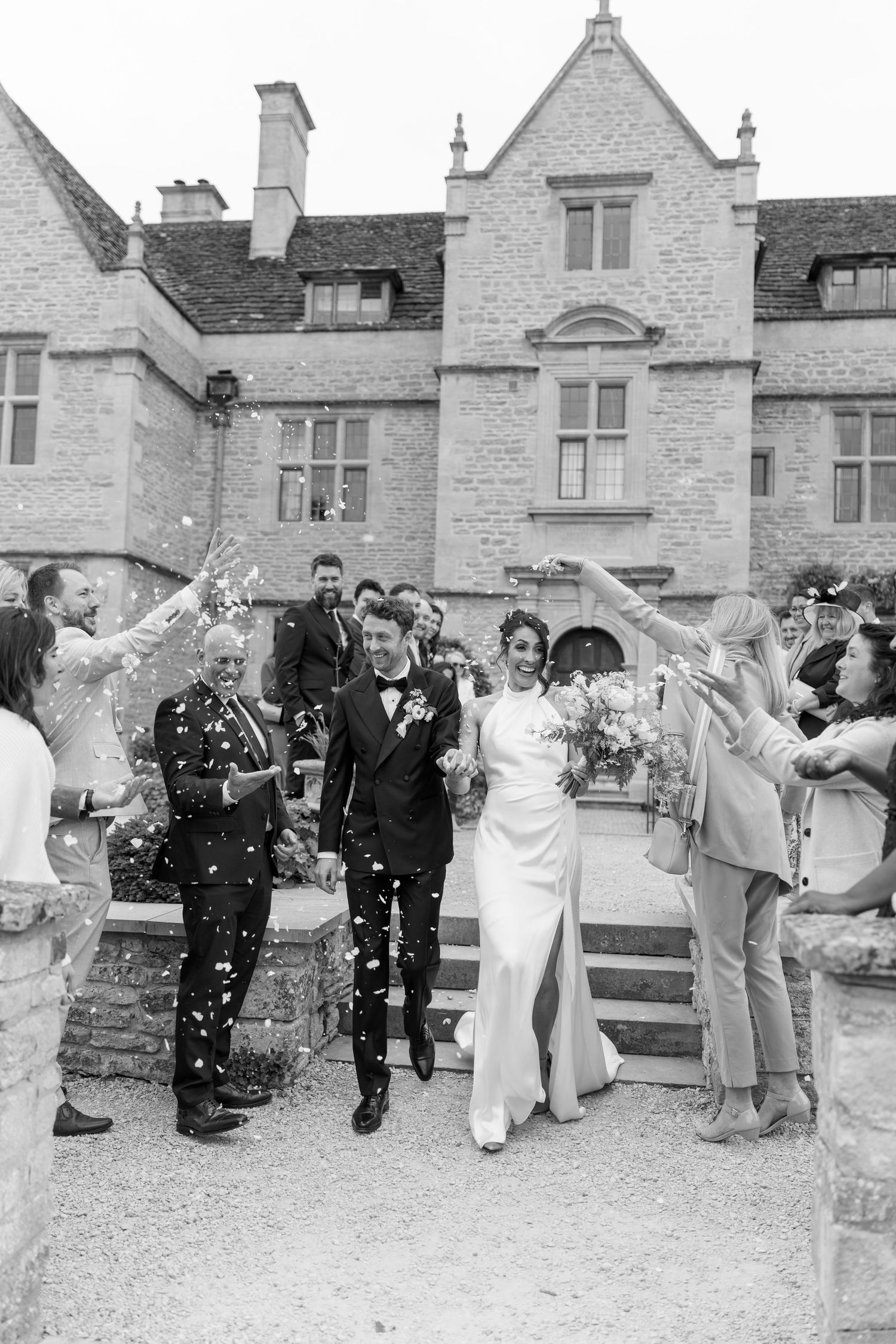 Black and white photo of newlyweds walking through confetti outside stone manor house.
