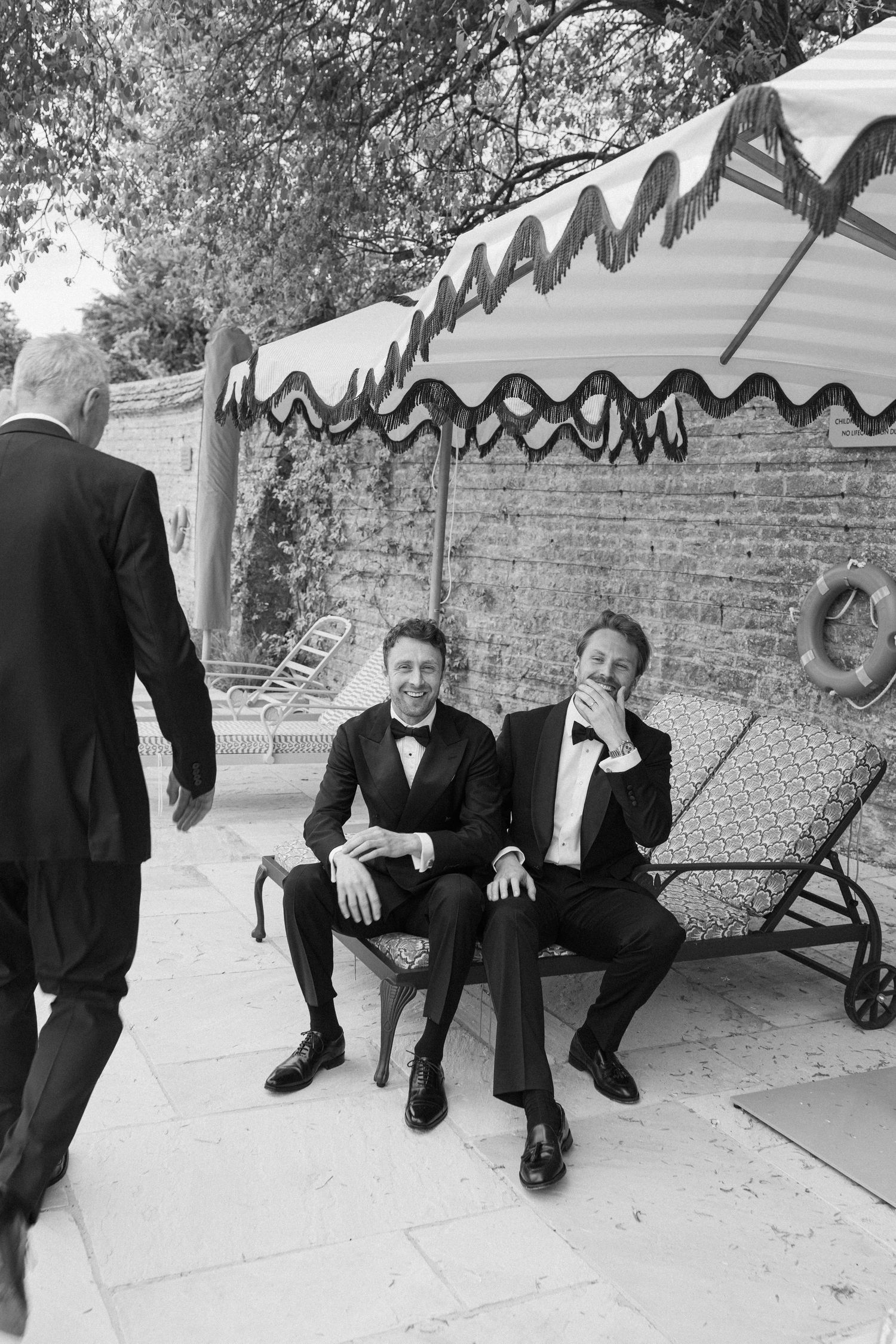 Two groomsmen in tuxedos sit under a striped canopy by a pool at an elegant outdoor wedding venue.