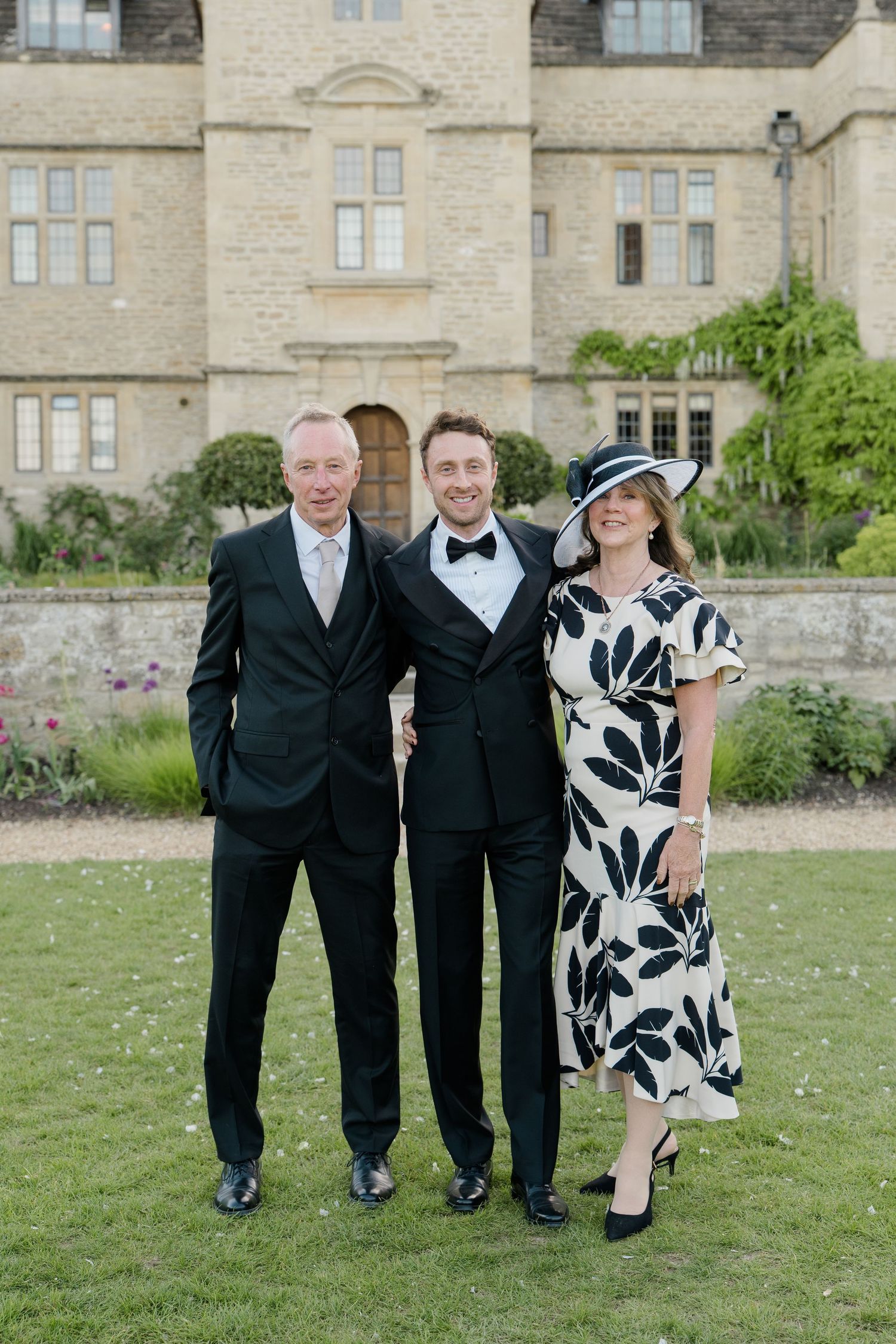 Groom in black tuxedo poses with parents outside stone manor, mother wearing floral dress and wide-brim hat.