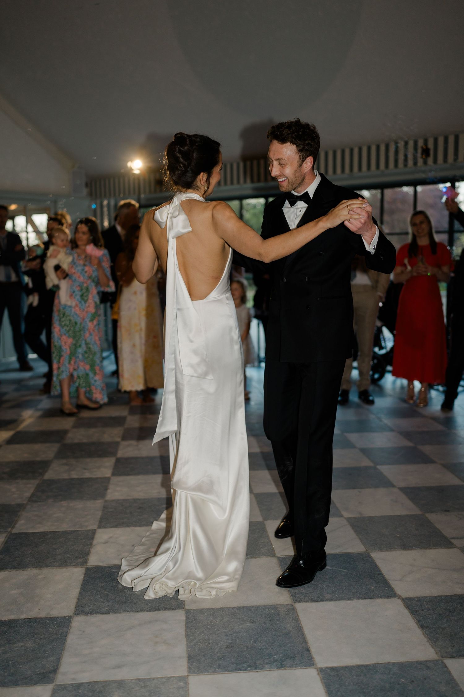 Bride in backless white satin gown shares first dance with groom in tuxedo on checkered ballroom floor.
