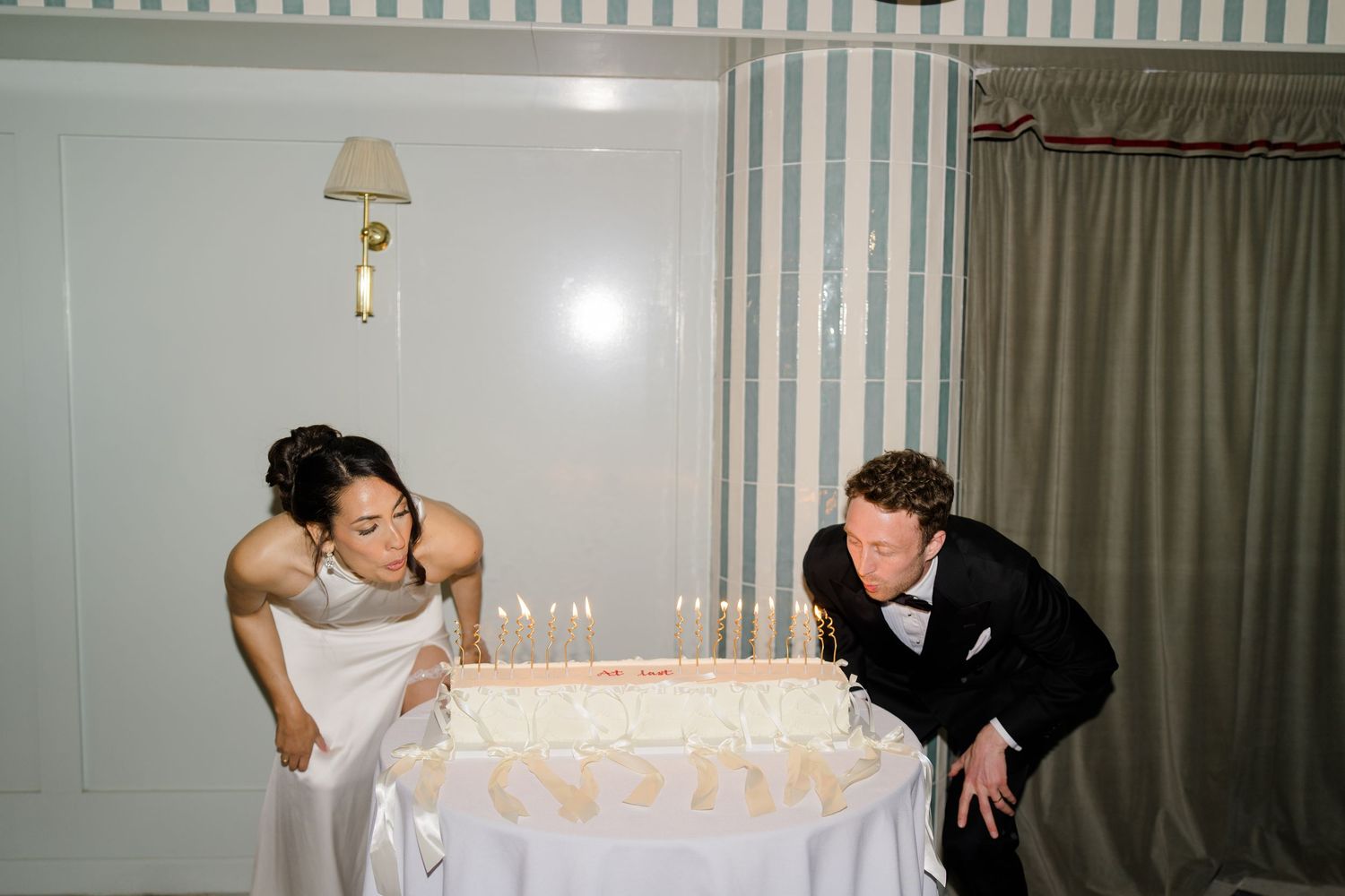 Bride and groom lean in together to blow out candles on elegant white wedding cake at reception.