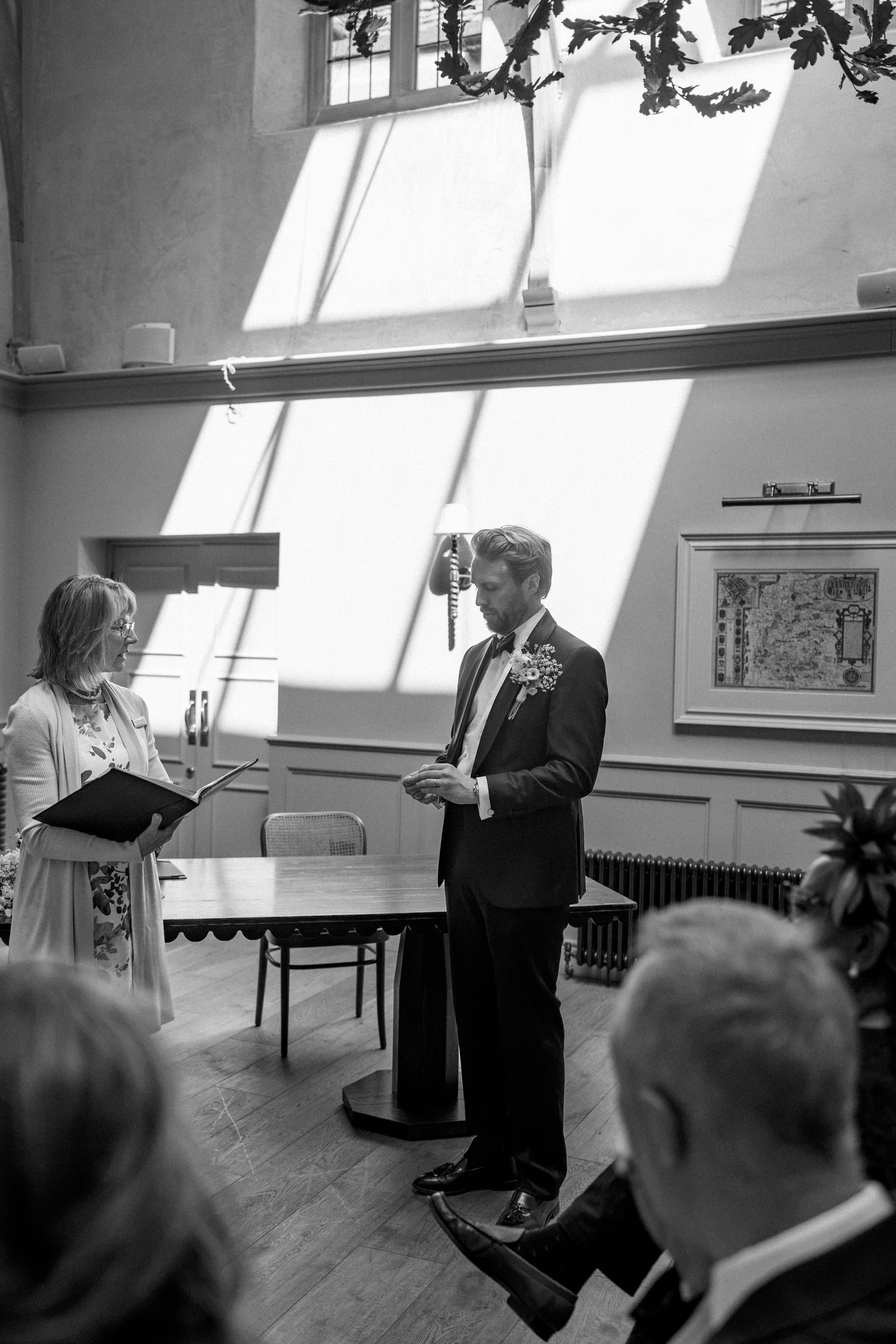 Groom reads vows at indoor wedding ceremony in black and white photo with sunlit windows.