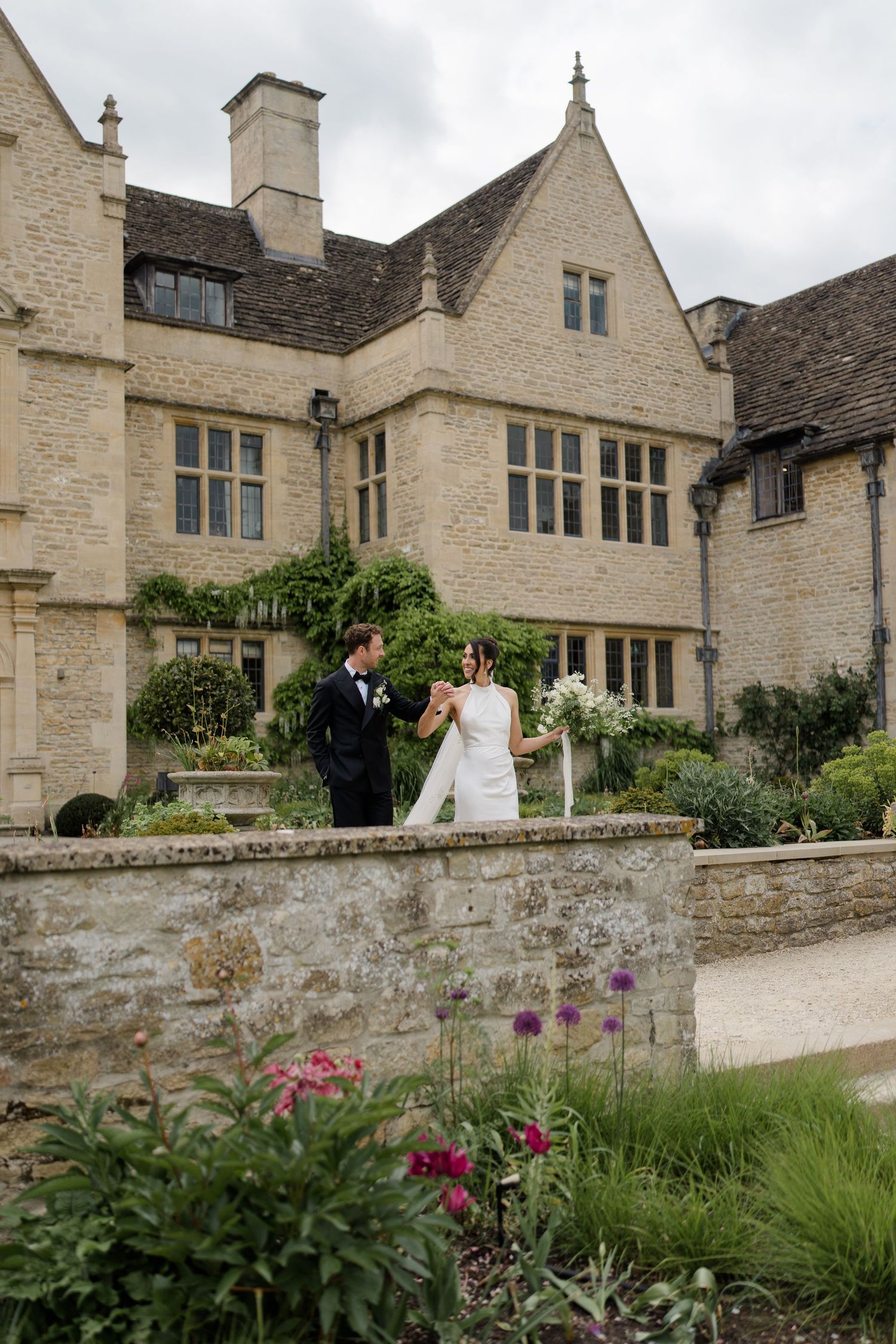 Bride and groom holding hands in a lush garden beside a historic Cotswolds stone country house.