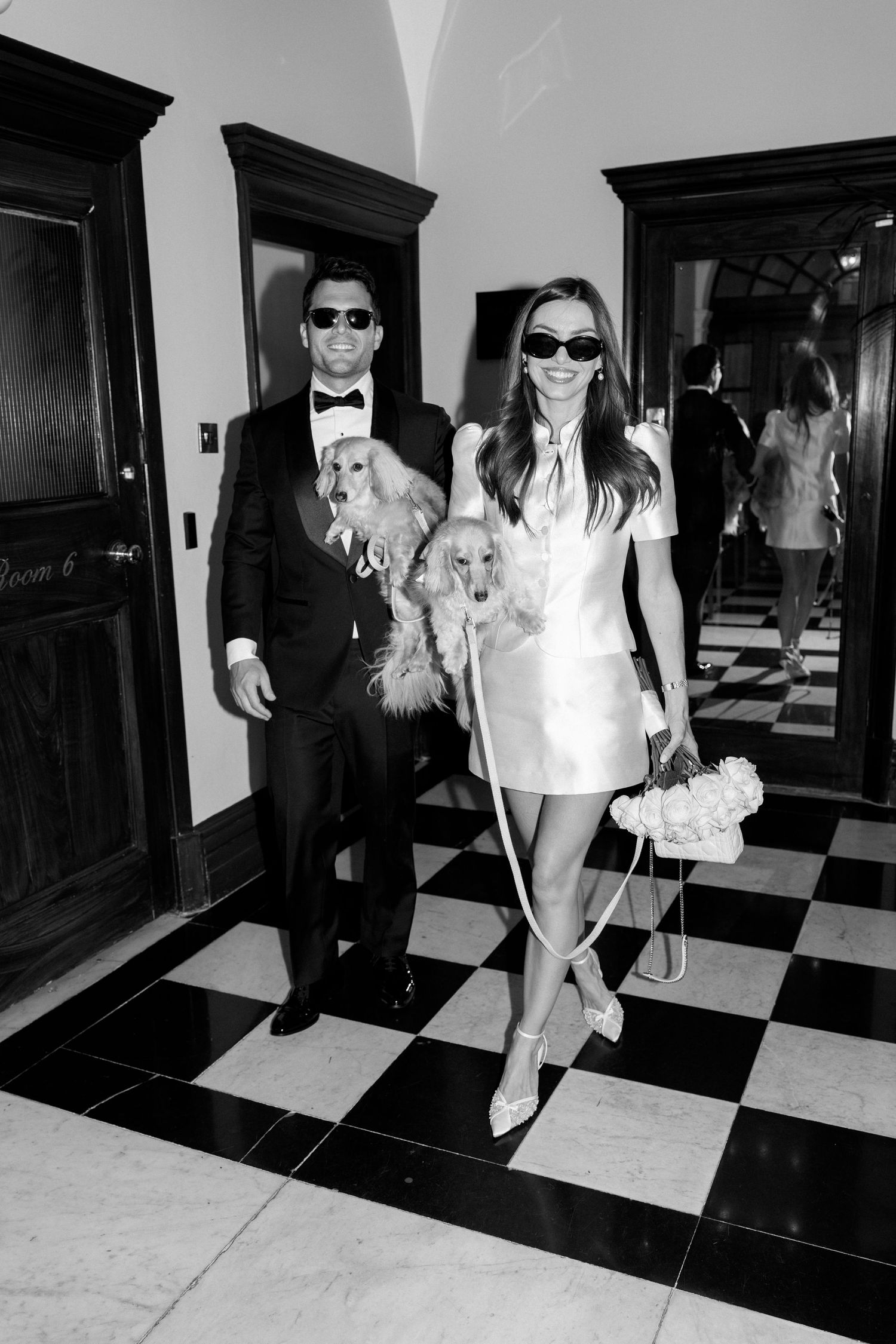 Elegant couple in formal attire with two fluffy dogs on checkered floor in black and white photo.