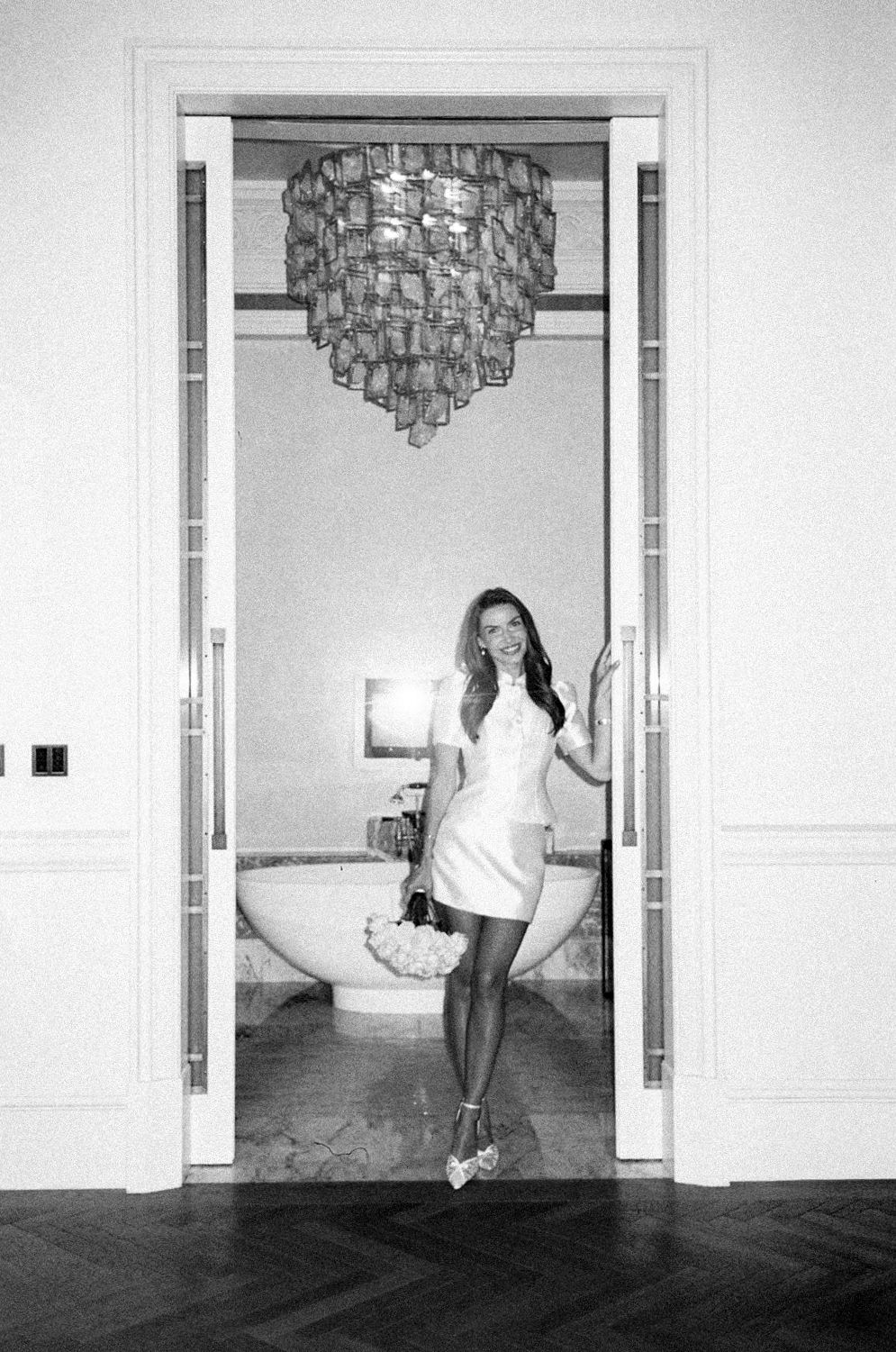 Woman in white mini dress posing in elegant doorway with chandelier and bathtub in black and white photo.
