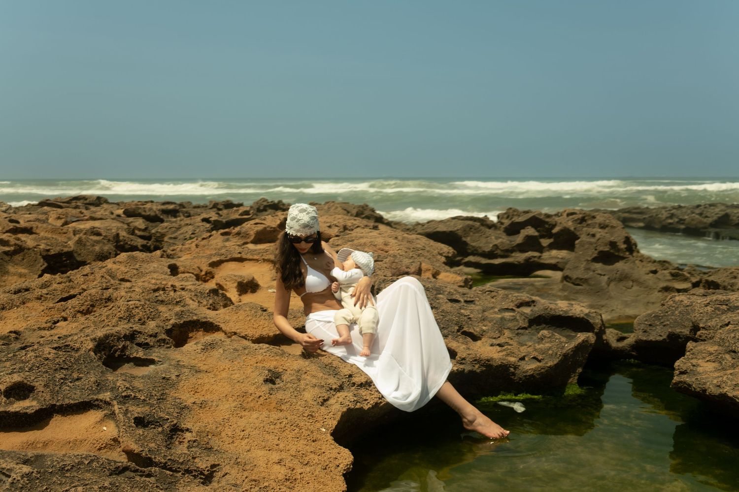 Two women in white outfits sit on rocky coastal shore with ocean waves in background.