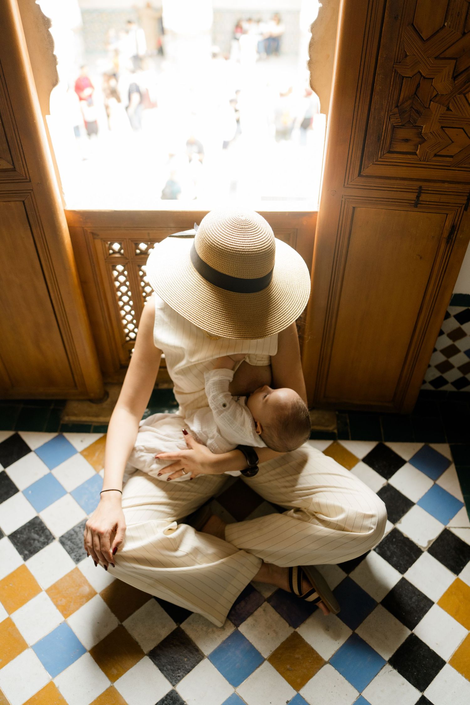 Mother in straw hat breastfeeding baby while sitting cross-legged on colorful diamond-patterned tile floor.