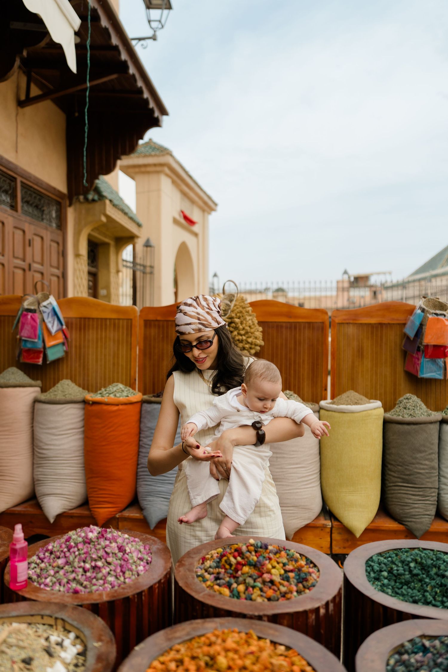 Woman holding baby browses colorful spices and herbs at a vibrant Moroccan market stall.