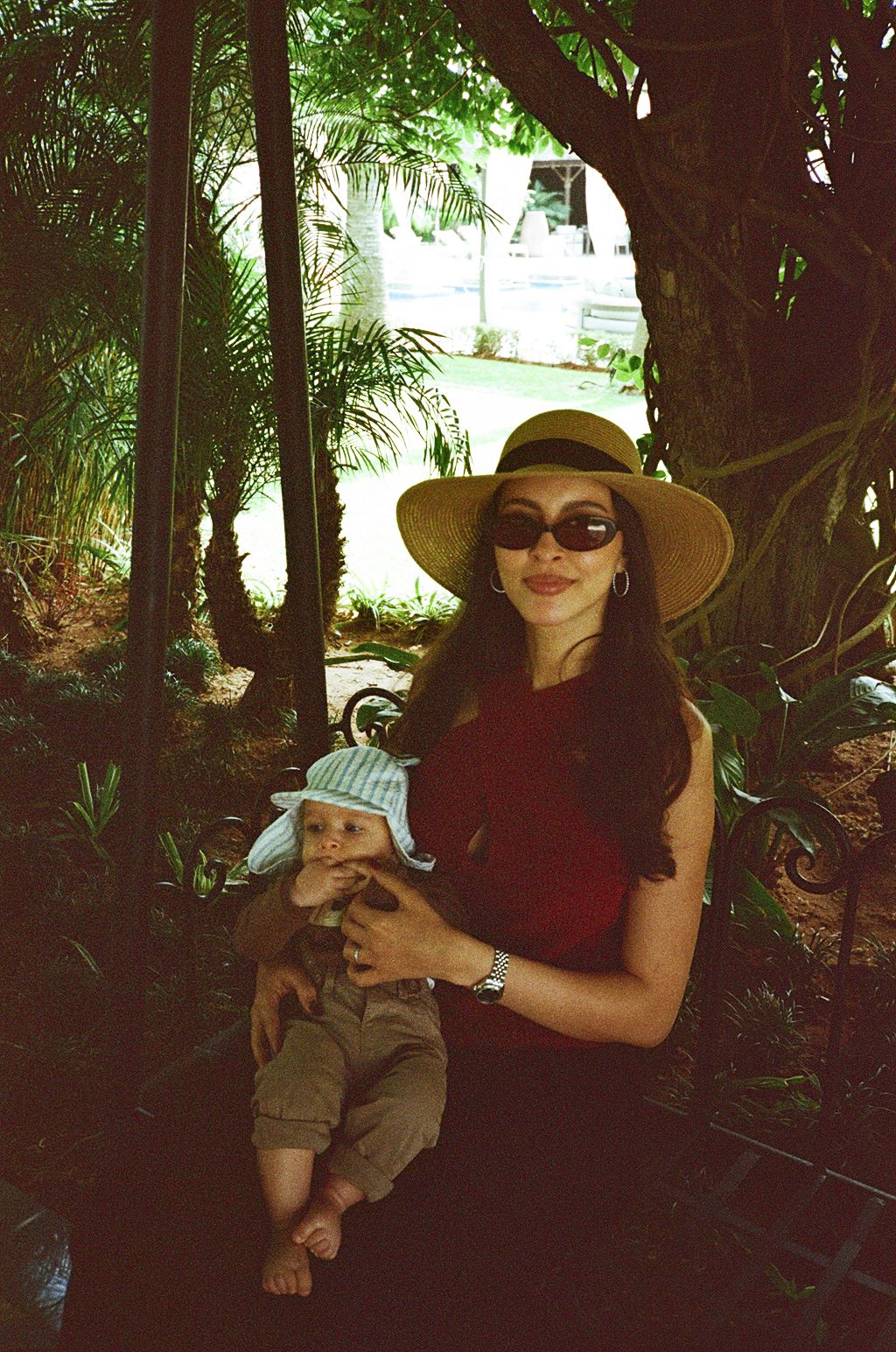 Woman in sun hat and sunglasses holding a baby in a lush garden setting.