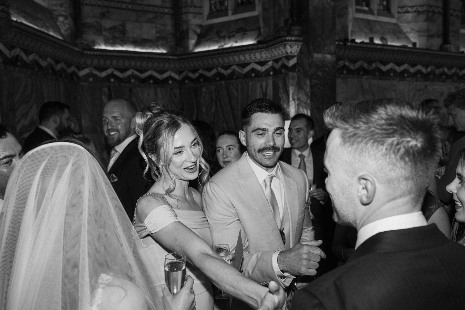 Black and white photo of wedding guests congratulating smiling couple inside ornate church venue.