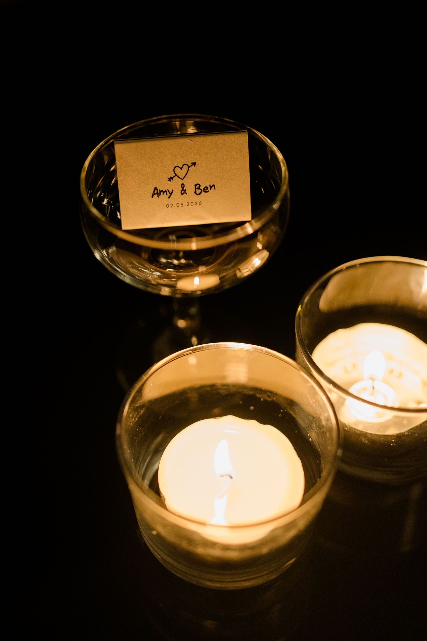 Romantic wedding candles in glass holders beside a card reading Amy and Ben on a dark surface.