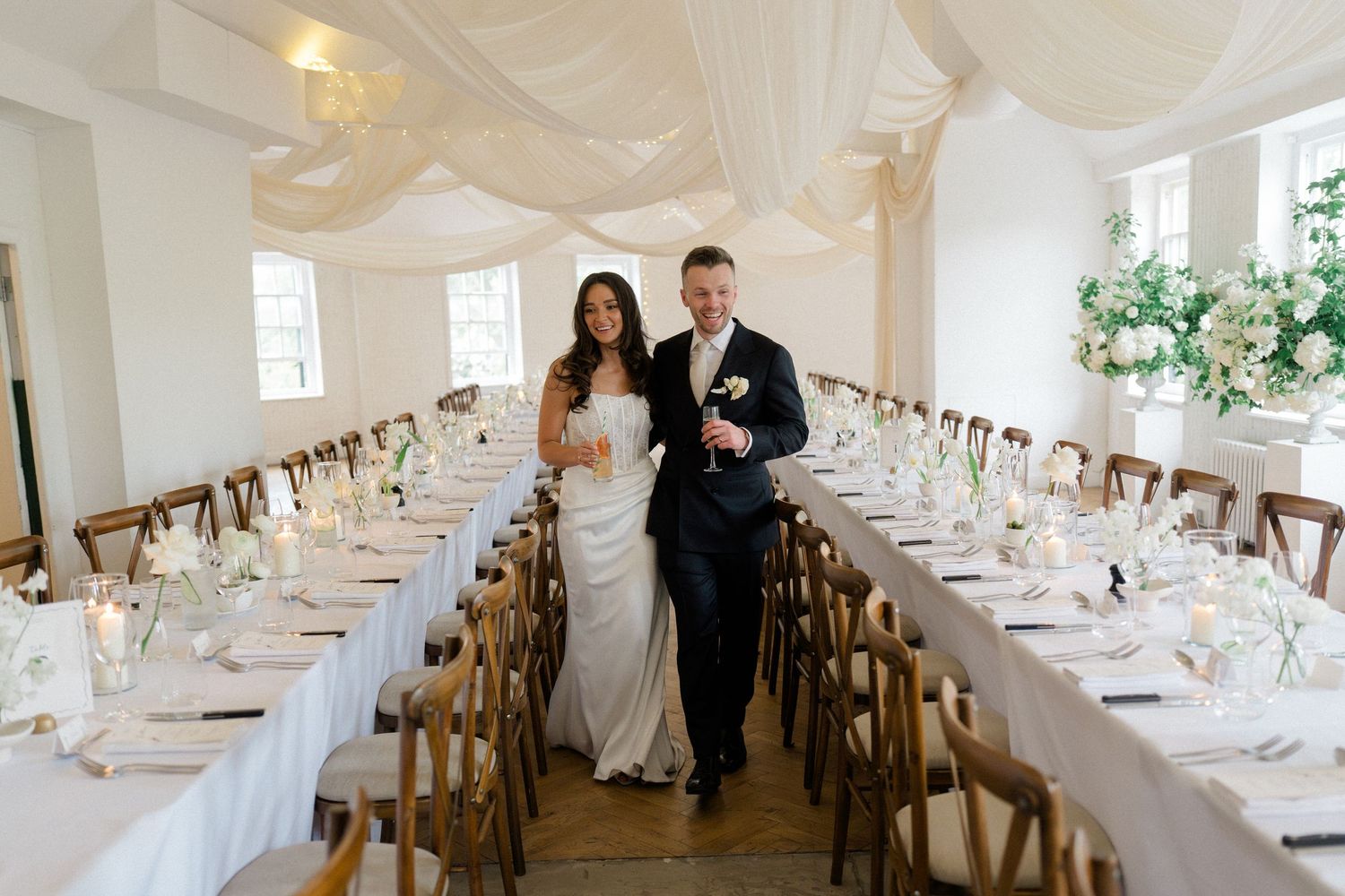 Bride and groom walking through an elegantly decorated wedding reception hall with long tables.